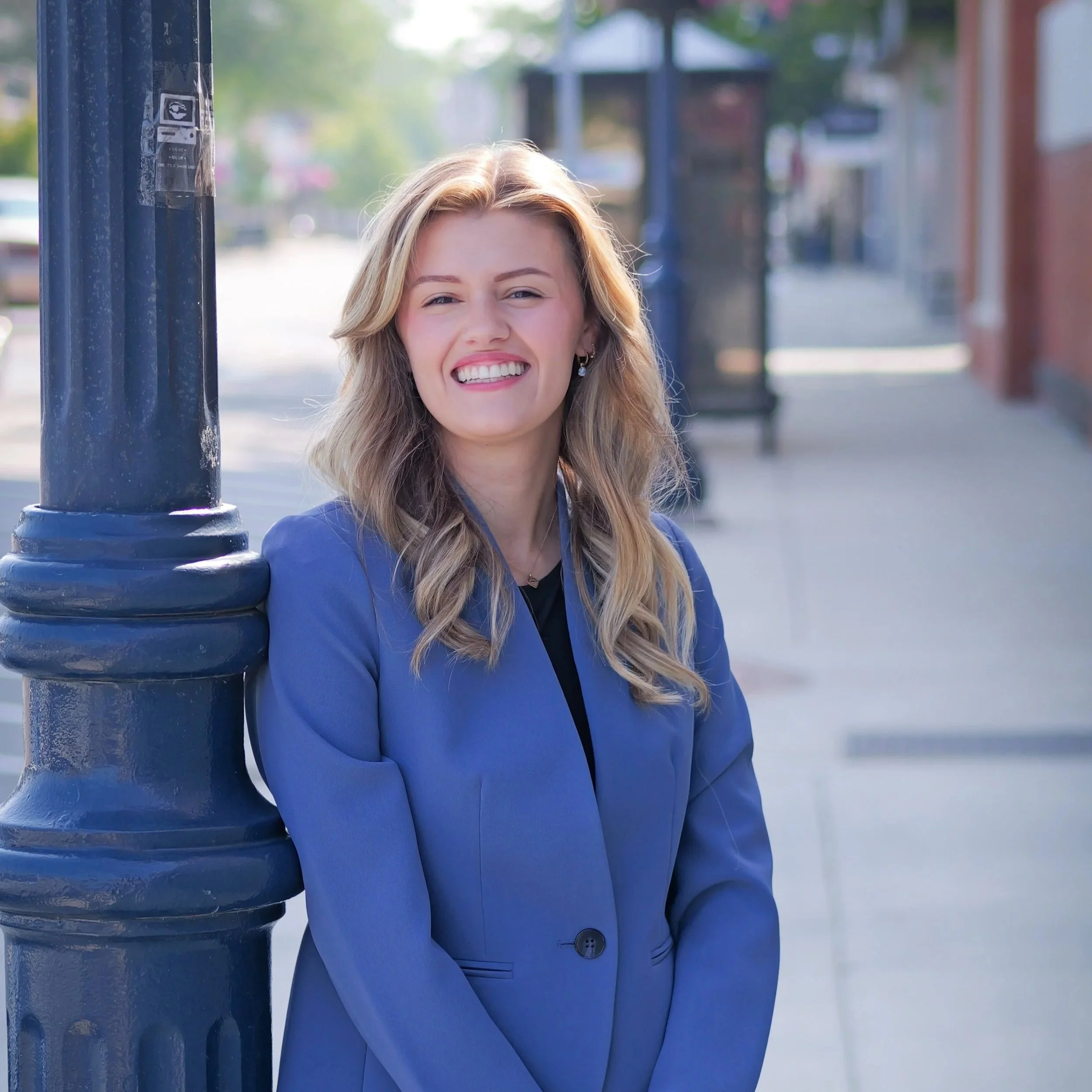 A smiling woman with blonde hair in a blue blazer standing on a city sidewalk near a black lamppost.