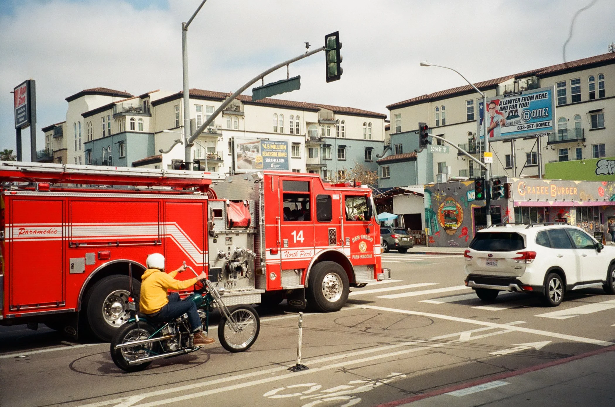 A red fire truck from San Diego North Park Fire-Rescue is stopped at an intersection. A person riding a motorcycle is passing near the fire truck. The street has marked bike lanes, and there are cars on the road. In the background, there are multi-story residential buildings, billboards, and a colorful restaurant called Grazee Burger on the corner.