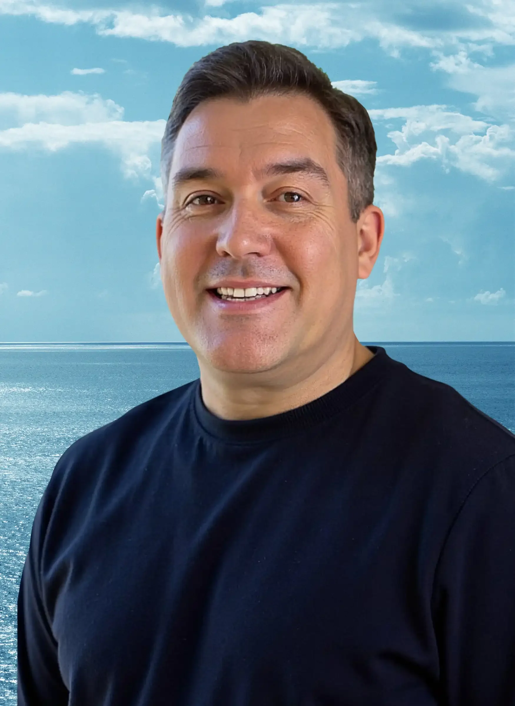 Close-up of a smiling man with short dark hair wearing a black shirt, with an ocean and cloudy sky in the background.