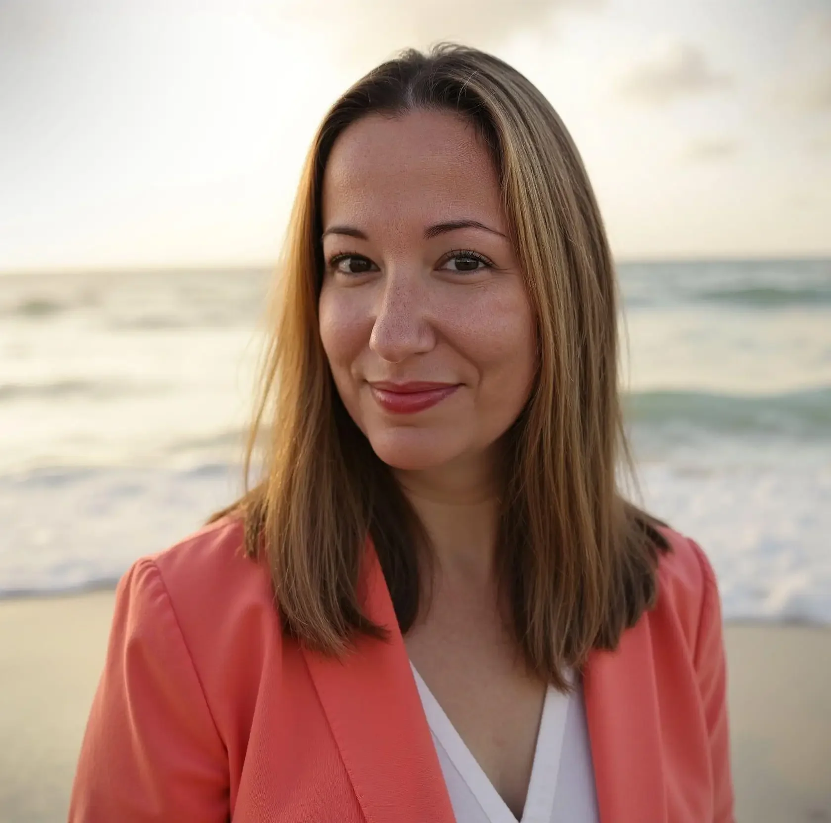 A woman with long brown hair smiling on a beach, wearing a coral blazer over a white top.