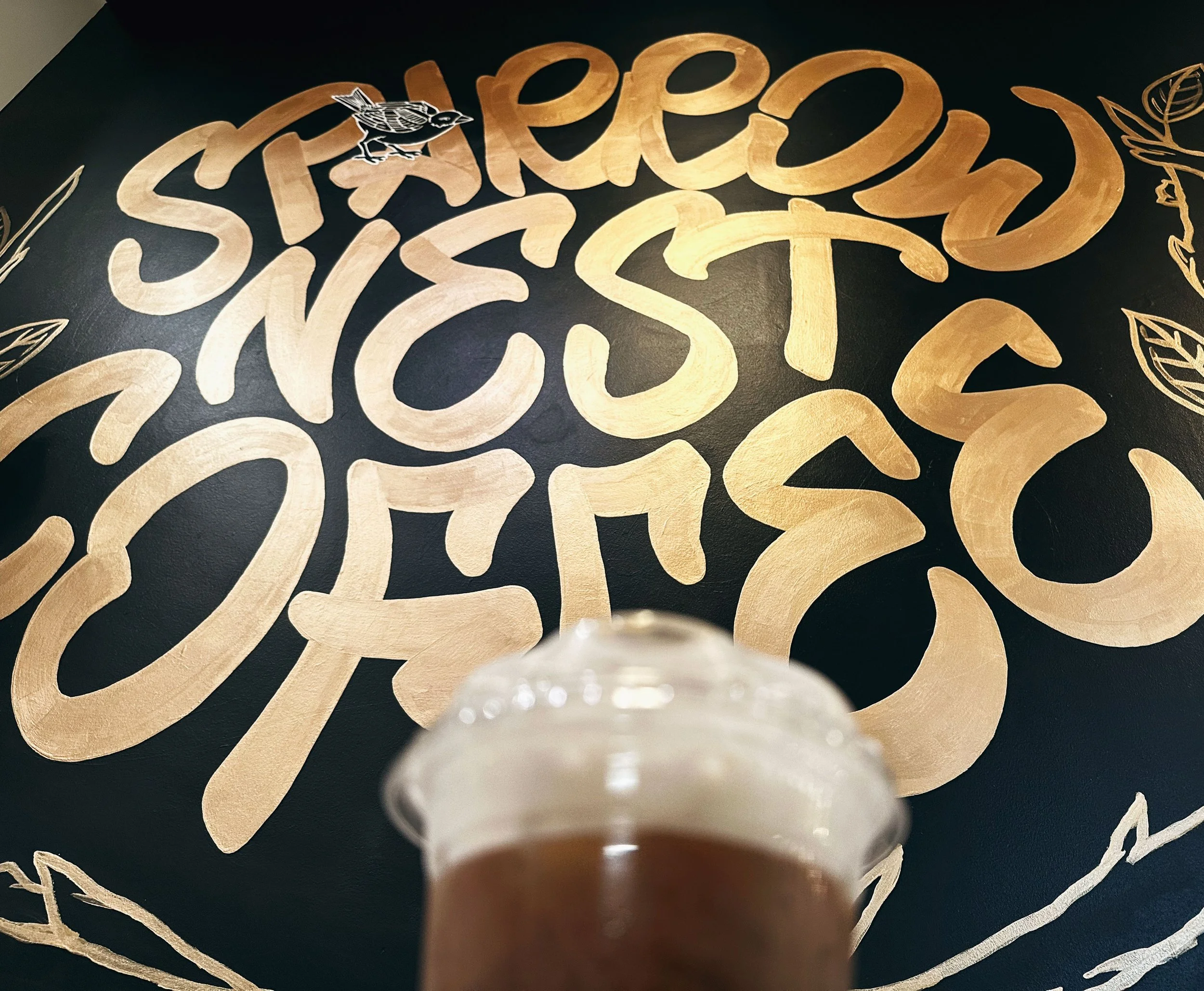 Completed Sparrow Nest Coffee hand-lettered mural on black wall with iced coffee in foreground