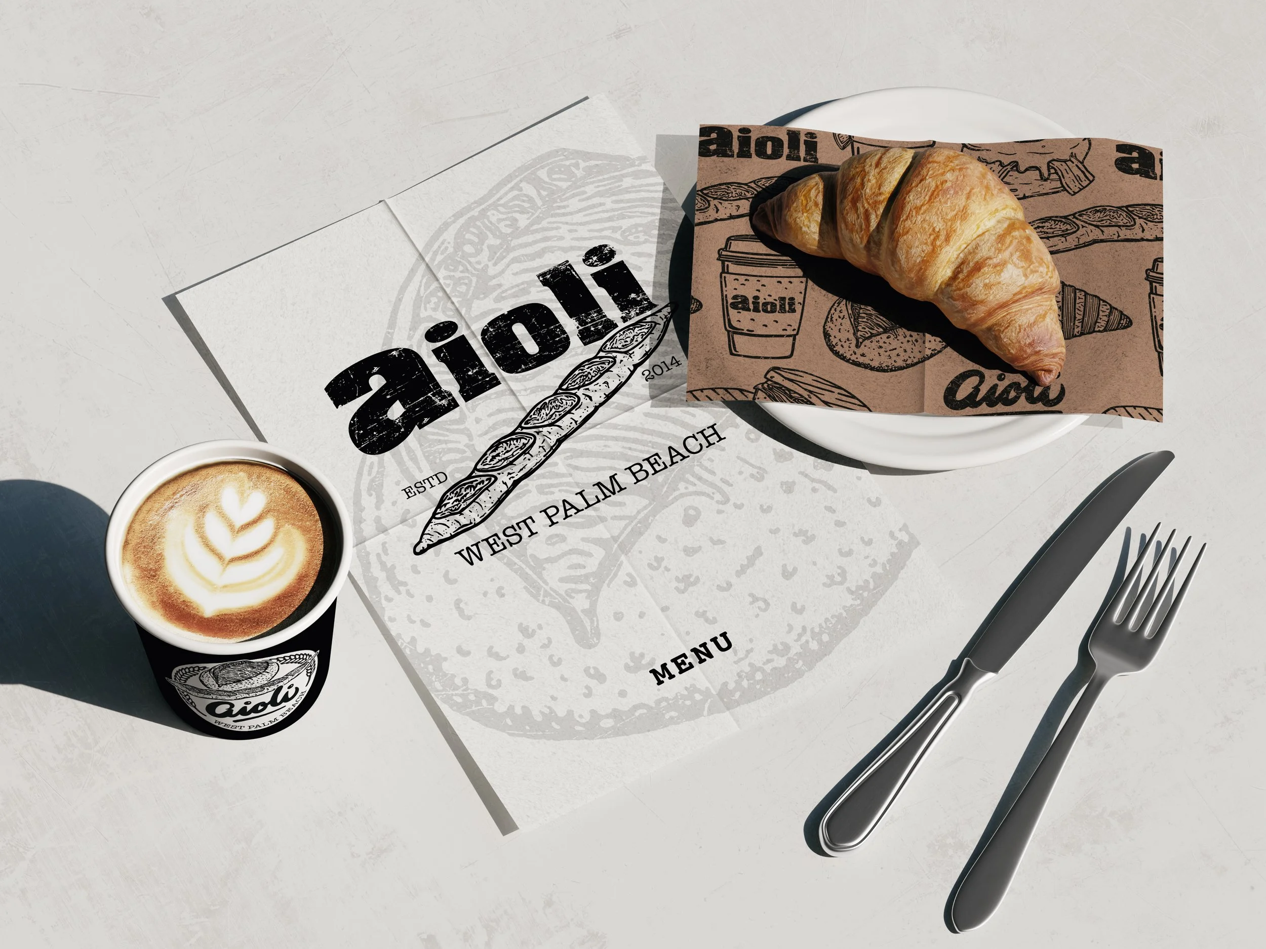 Aioli branded table setting featuring illustrated deli paper, menu, coffee cup with latte art, fresh croissant, and utensils on marble surface