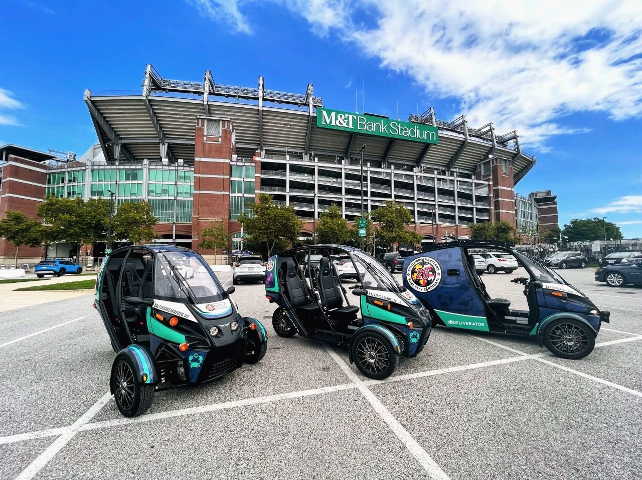 Three Arcimoto vehicles including FUVs and Deliverator parked outside stadium for Baltimore 2026 FIFA World Cup Host City bid