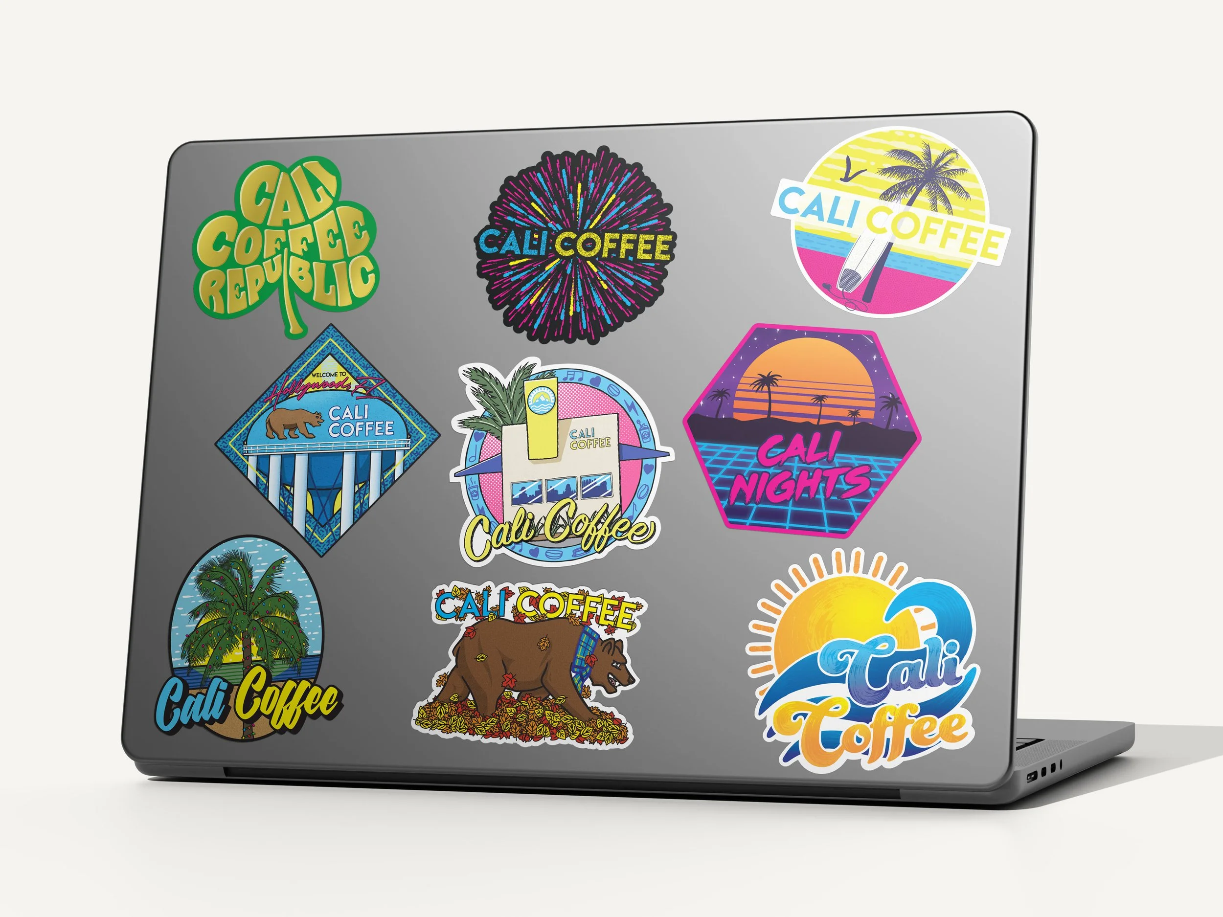 Collection of Cali Coffee monthly stickers on laptop showing 2019 series themes - St. Patrick's Day shamrock, fireworks, outrun aesthetics, palm tree with Christmas lights, and seasonal designs