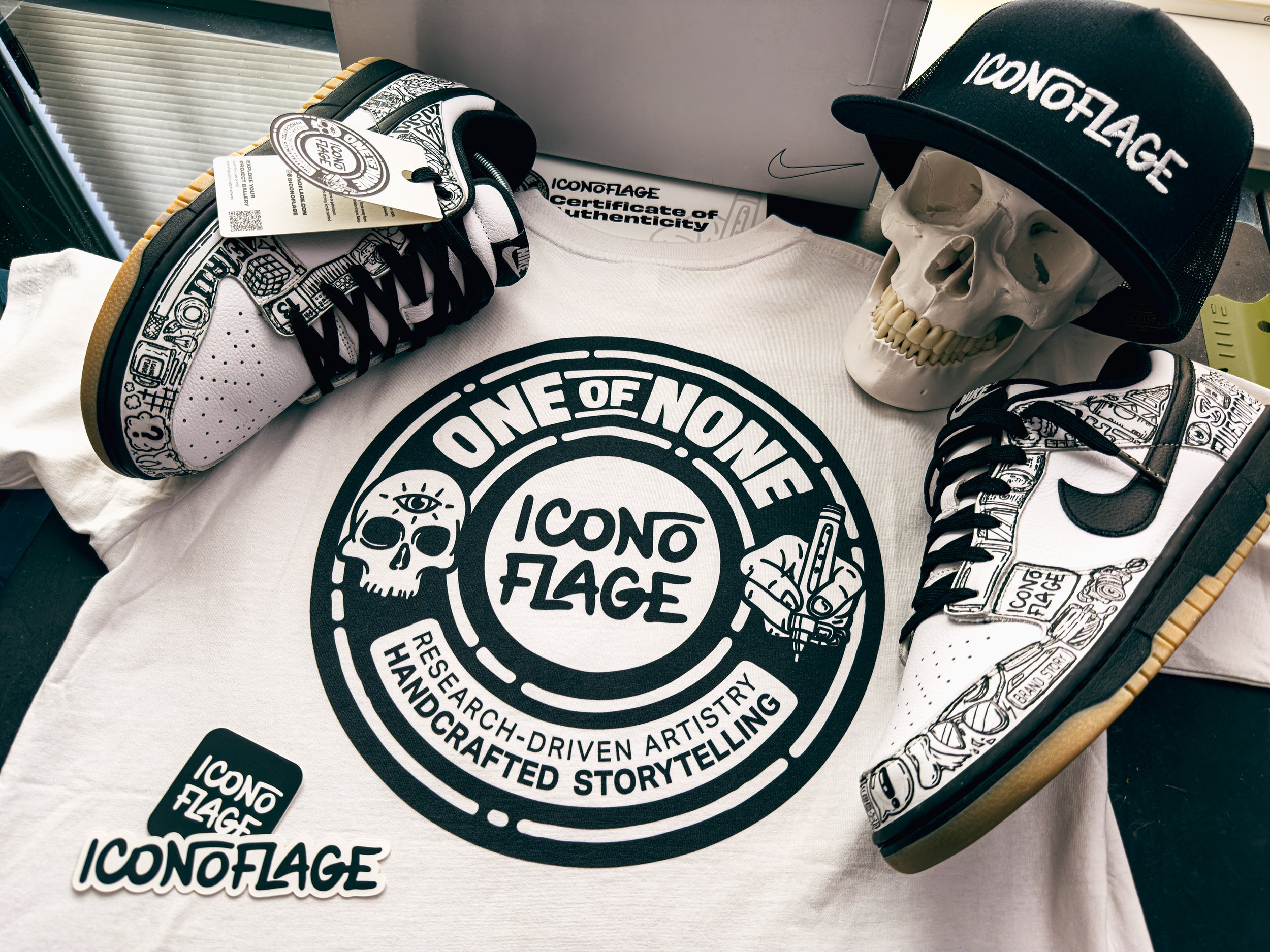 Meta II Iconoflage Nike Dunks with custom packaging including certificate of authenticity, hangtag, branded hat, skull prop, and white t-shirt featuring One of None seal