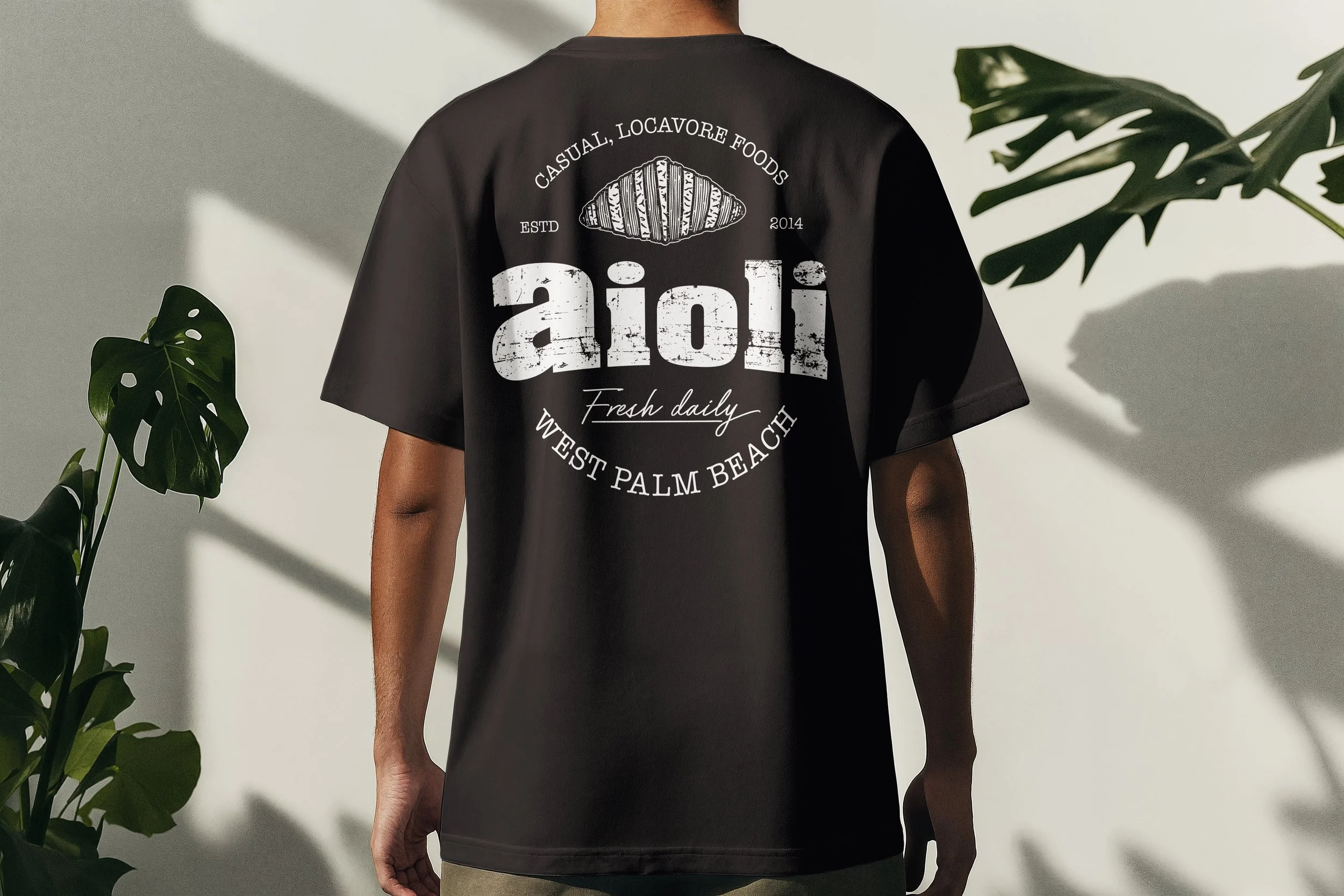 Back view of black Aioli t-shirt featuring large logo with illustrated croissant design and West Palm Beach location text