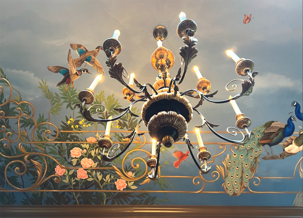 Ornate chandelier with multiple lights hanging from a ceiling decorated with painted peacocks, birds, and floral designs.