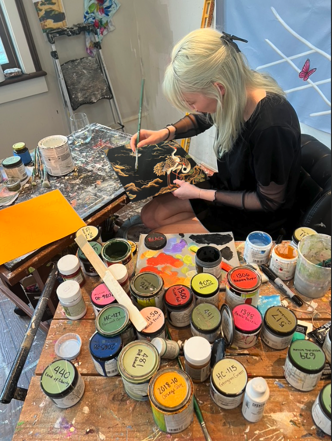 with platinum blonde hair, seated at a cluttered wooden desk, is painting a detailed design on a black canvas with gold and white accents. The desk is filled with various jars of paint, brushes, and art supplies, and she is focused on her work in a bright studio space.