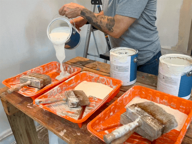Person pouring white paint or primer into orange paint trays with large paintbrushes resting inside, on a wooden table.
