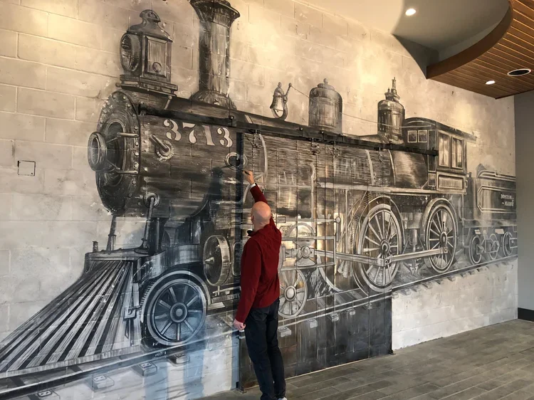 Andrew Tedesco painting a large mural of a vintage steam locomotive on a brick wall at The Archer Hotel in Massachusetts.