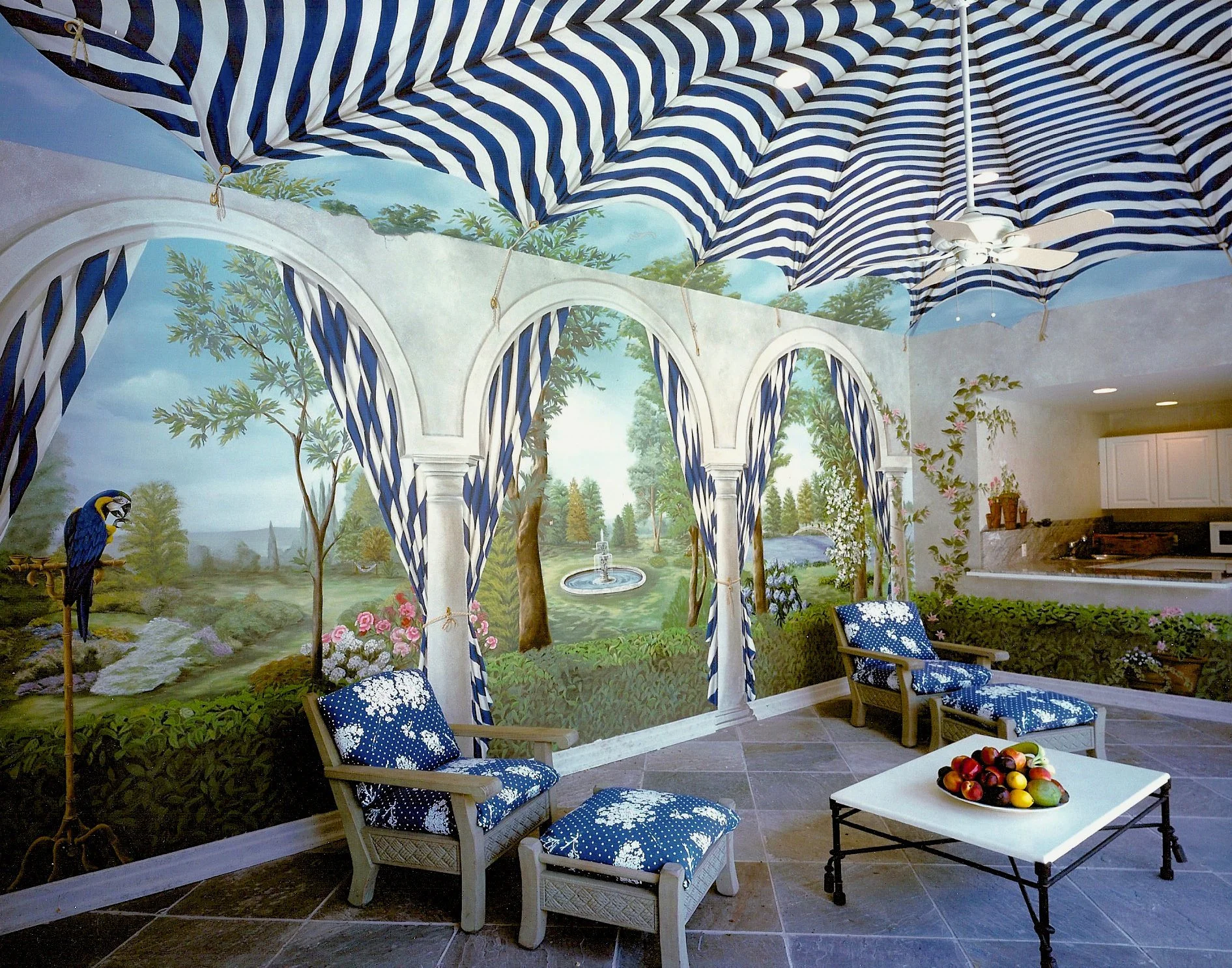 Indoor living area with hand-painted mural of a scenic landscape featuring trees, a fountain, and a parrot, along with blue and white striped curtains and ceiling, two armchairs with matching cushions, a small white table with a bowl of assorted frui