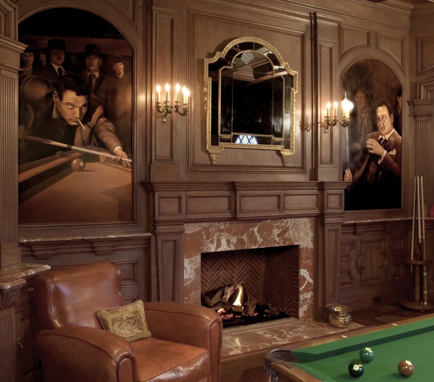 A cozy room with a wooden-paneled wall, a fireplace with fire burning, a brown leather chair with a decorative pillow, and a pool table with green felt and billiard balls. Two large wall paintings depict men playing snooker or pool.
