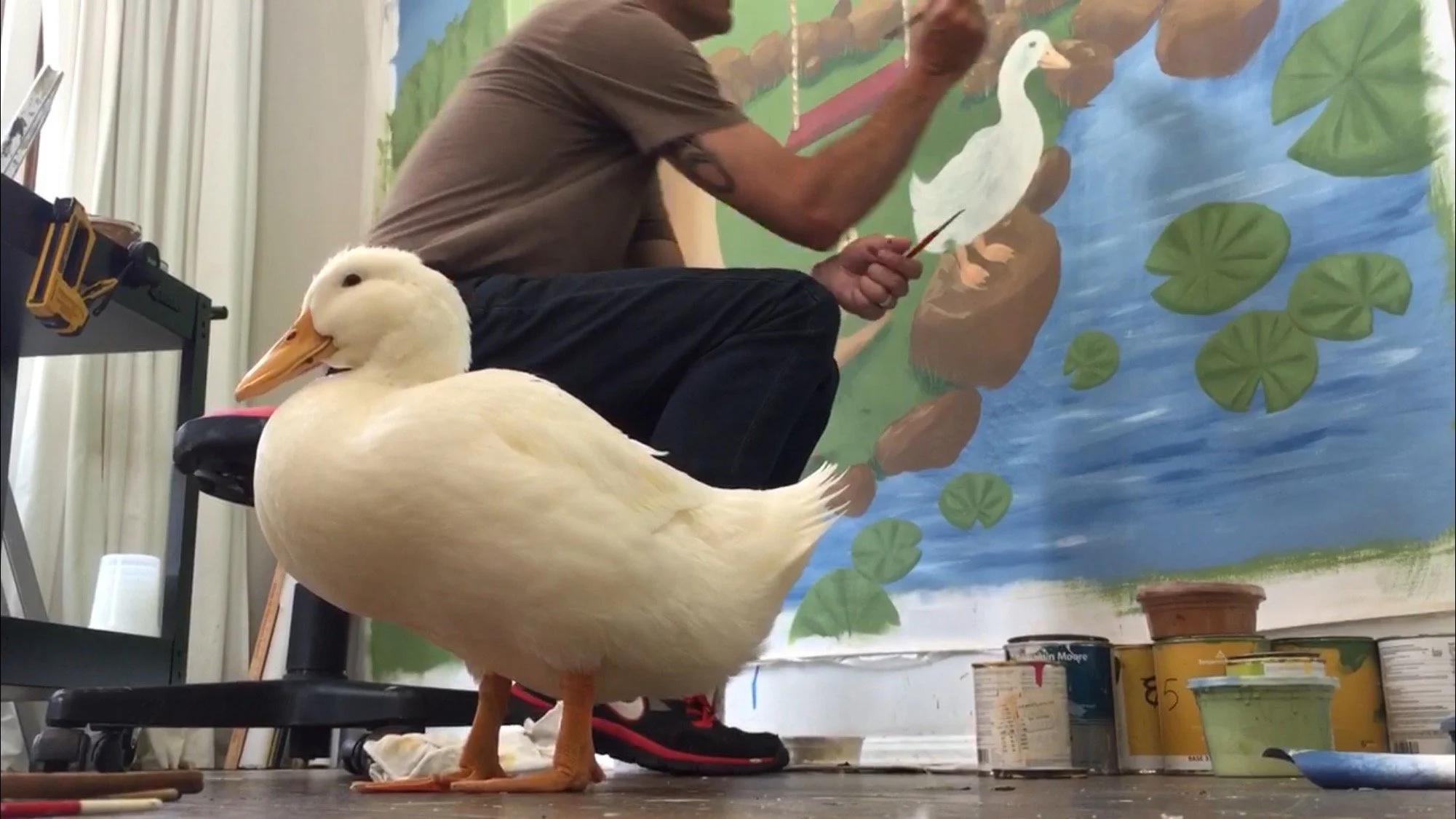 A white duck with orange beak and feet standing on the floor in front of a colorful pond mural, with a person painting the mural in the background.