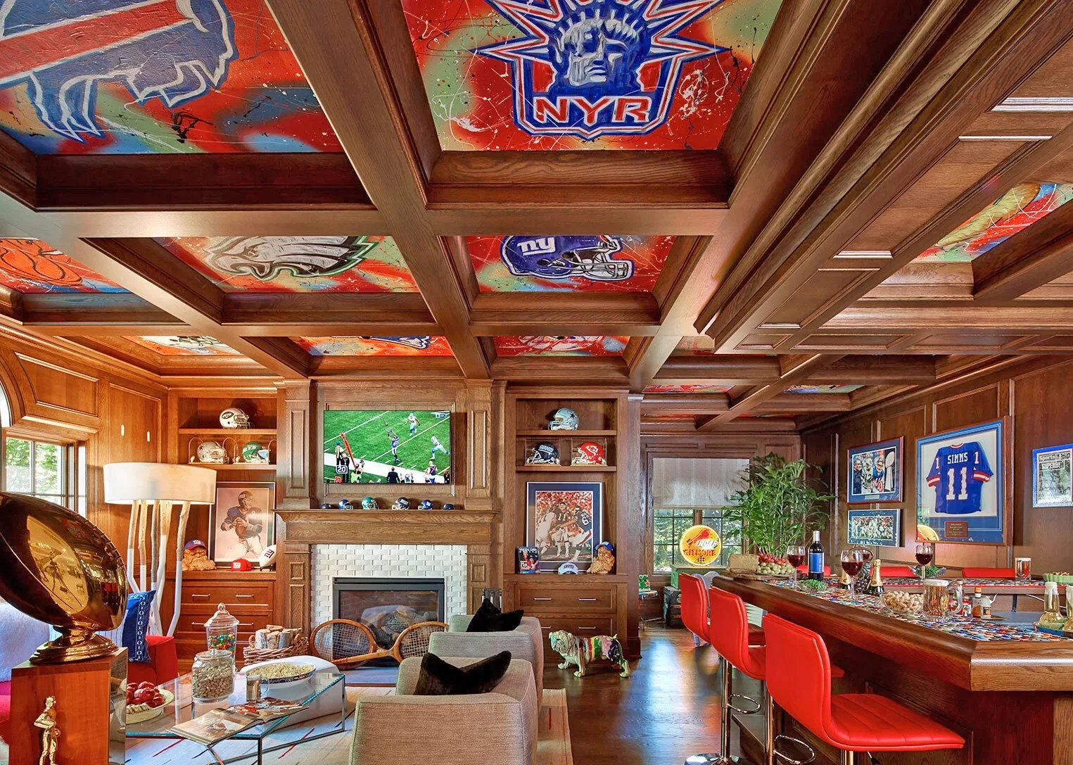 A sports-themed living room by Barbara Ostrom Associates with wooden walls, ceiling, and furniture, featuring NFL team logos on the ceiling, framed sports memorabilia, a flat-screen TV showing football, and a bar area with red chairs.
