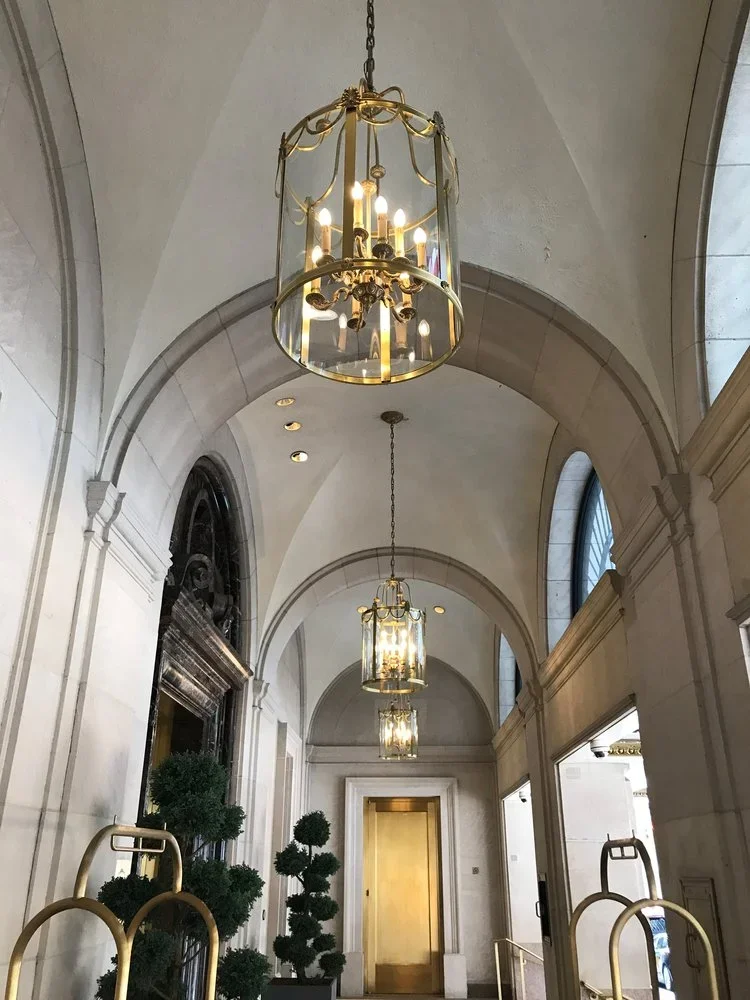 Elegant hotel lobby or corridor with high arched ceilings, hanging chandeliers, and decorative plants.