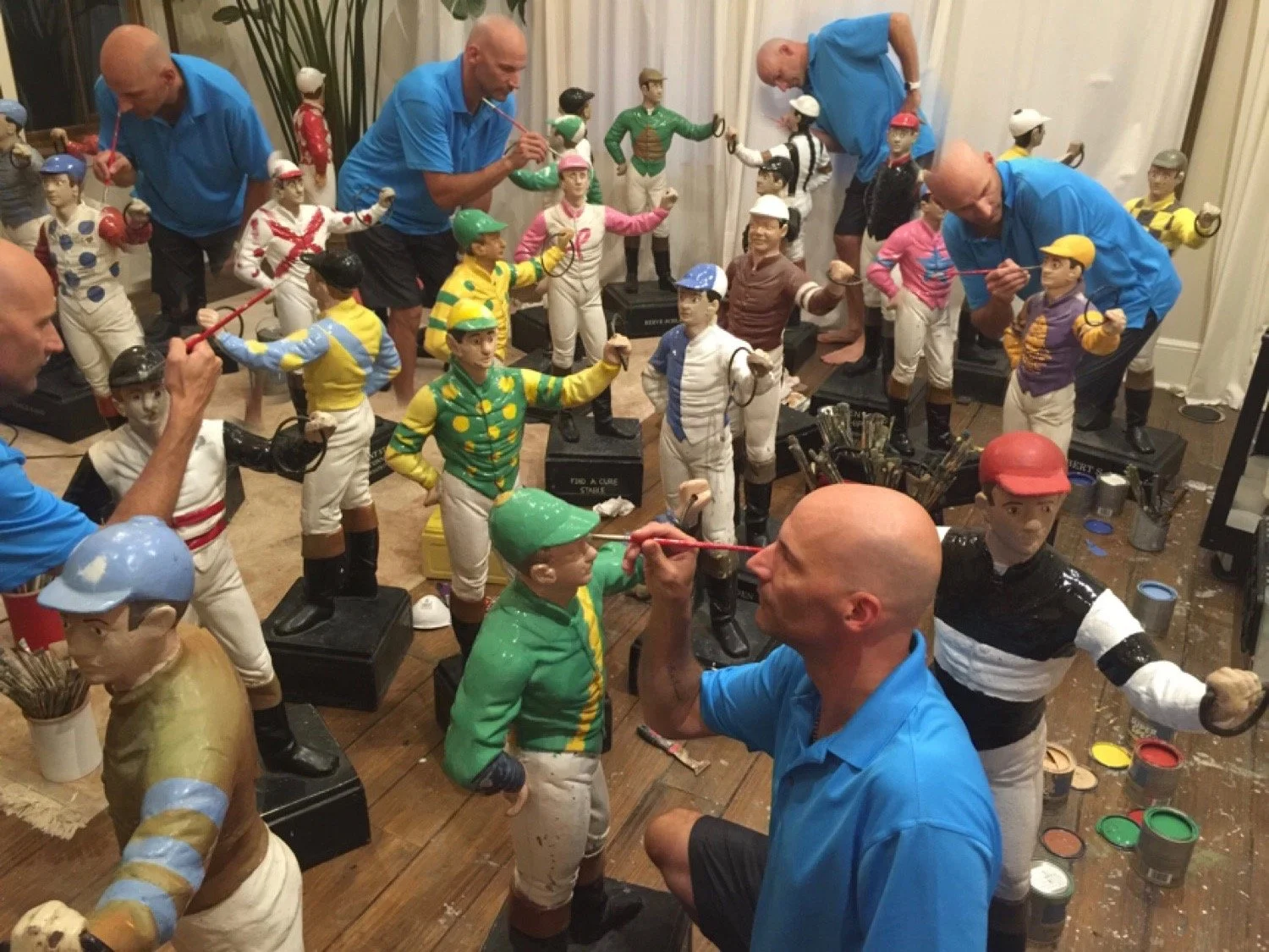 A group of men painting and decorating multiple jockey statue sculptures inside a room with wooden flooring and light-colored curtains.