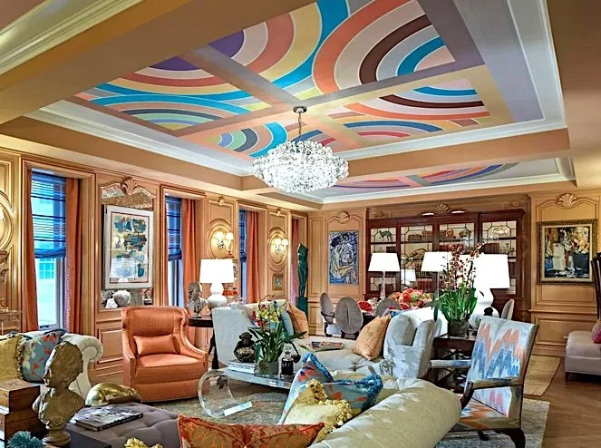 A luxurious living room with colorful painted ceiling, chandelier, beige walls, large windows with orange curtains, various sofas and armchairs, artwork on the walls, and decorative objects on tables and shelves.