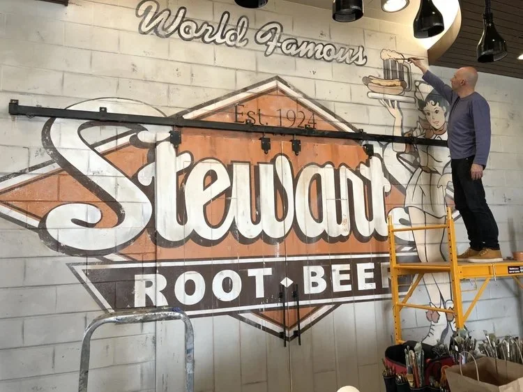 Andrew Tedesco standing on scaffolding painting a mural of a woman holding a tray with a burger and soda on a wall at Stewart Root Beer restaurant, with the text 'World Famous' above and the Stewart logo below at The Archer Hotel in New Jersey.