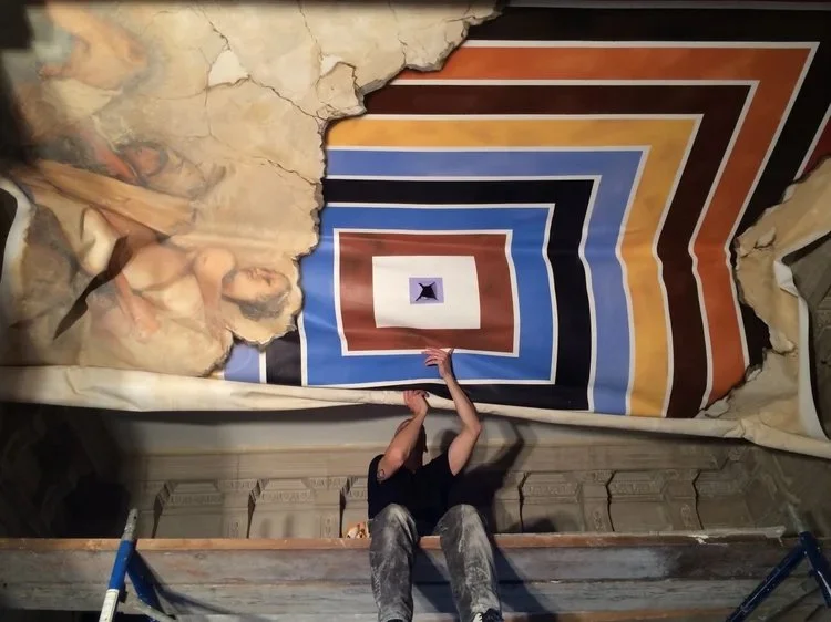 Person installing or repairing a ceiling mural that features a geometric pattern of colorful squares radiating outward, with some of the surrounding paper torn away.