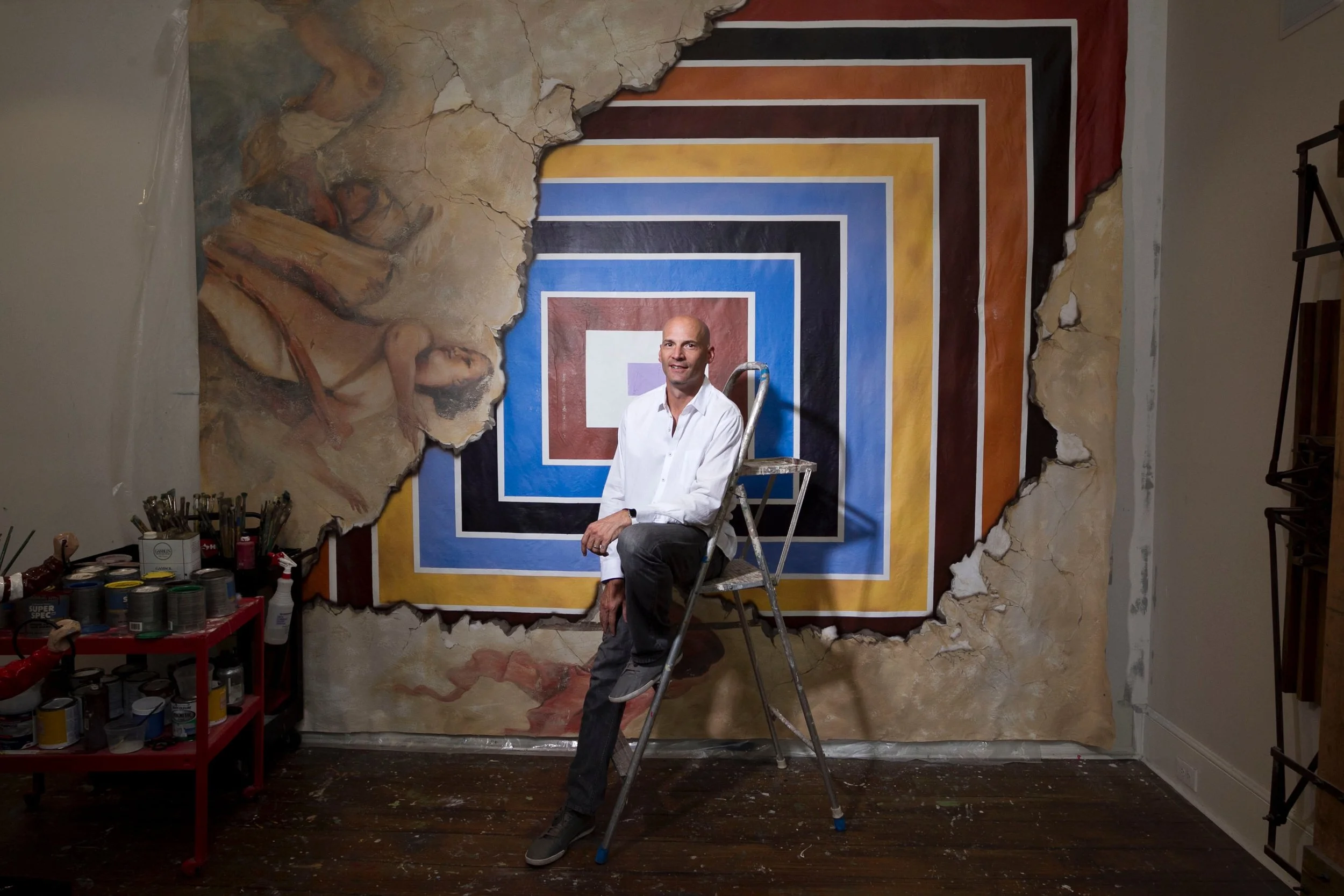An artist sits on a ladder in front of a wall with a geometric pattern of concentric squares in multiple colors; part of the wall has a mural that appears to be peeling away.