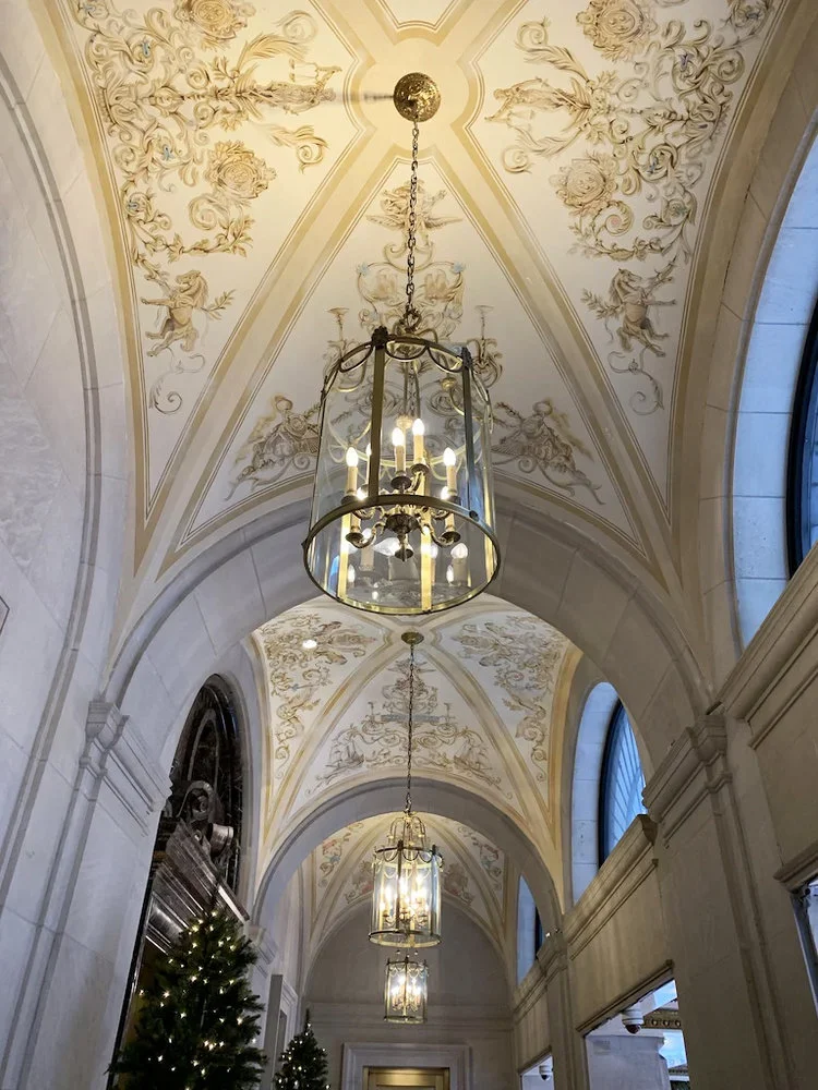 Decorative ceiling with floral and angel motifs, ornate chandeliers hanging from chains, arched windows along a corridor, decorated Christmas trees.