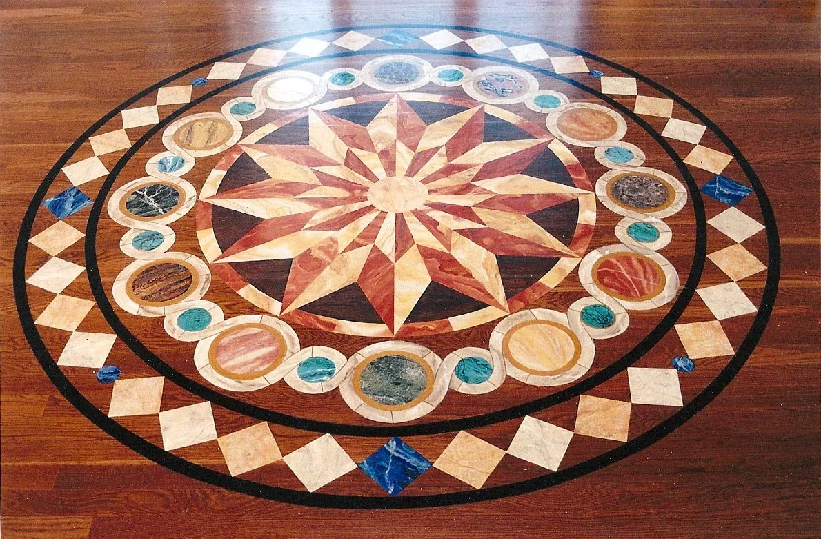 Intricate inlaid painted floor design of a geometric pattern on a wooden floor. The design features a central star pattern surrounded by circles with various colored stones, bordered by diamond shapes and concentric rings.