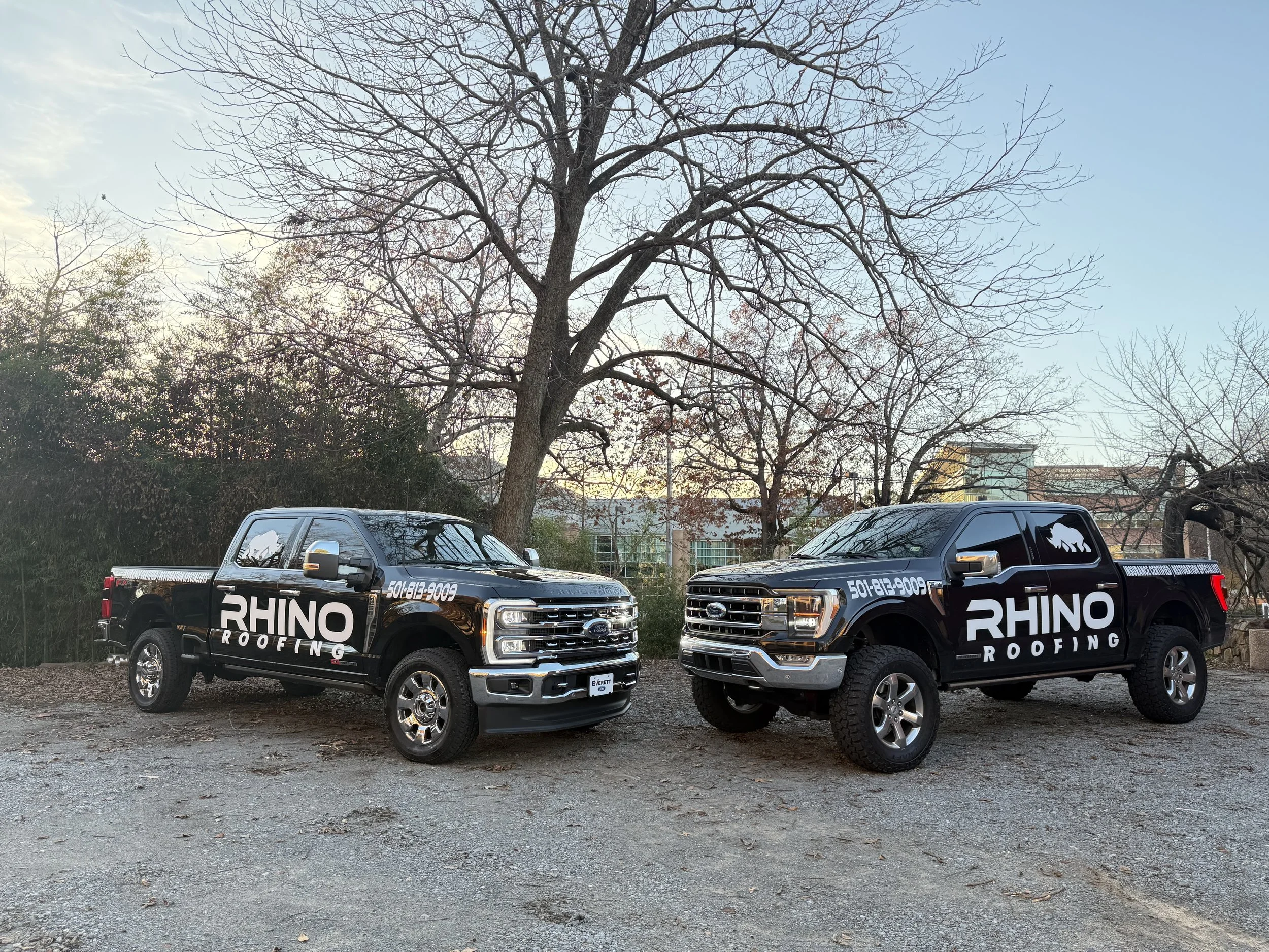 Two black pickup trucks with "RHINO ROOFING" branding on their sides are parked on a gravel surface, with leafless trees and a building in the background.