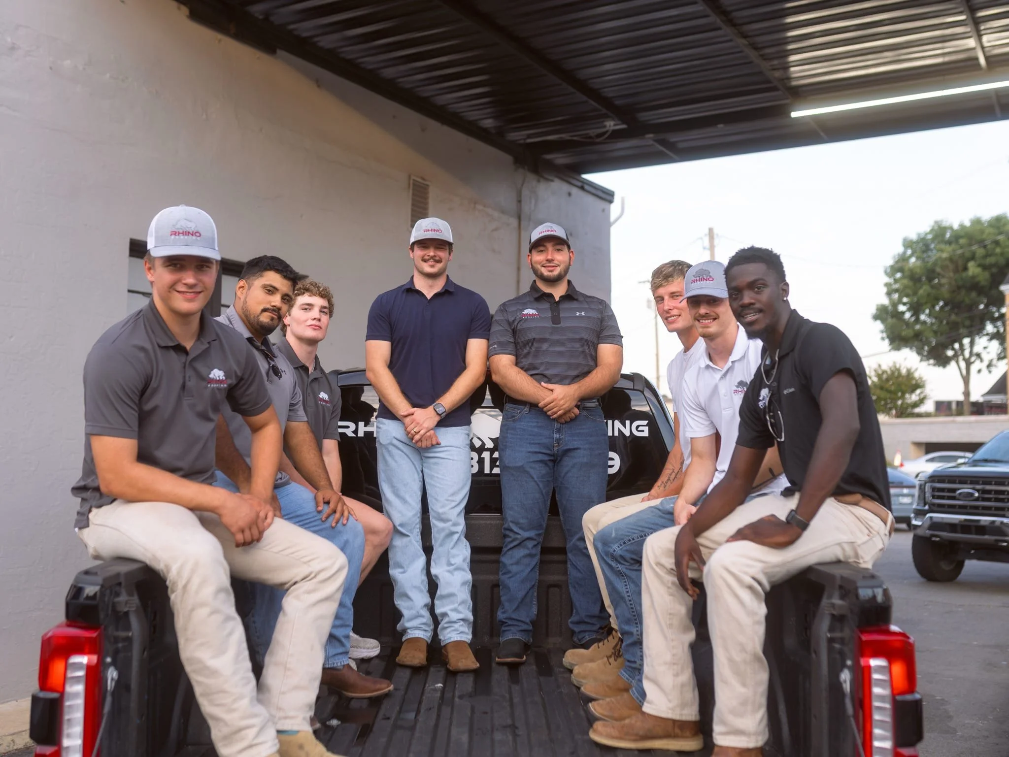 Group of eight young men, some wearing hats and polo shirts with a company logo, sitting and standing on the back of a black pickup truck parked outdoors, with trees and other vehicles in the background.