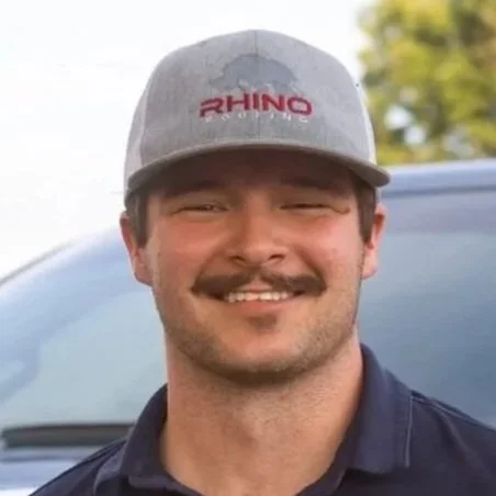 A man with a mustache smiling, wearing a gray baseball cap with 'RHINO' written on it, and a navy blue polo shirt, standing outdoors.