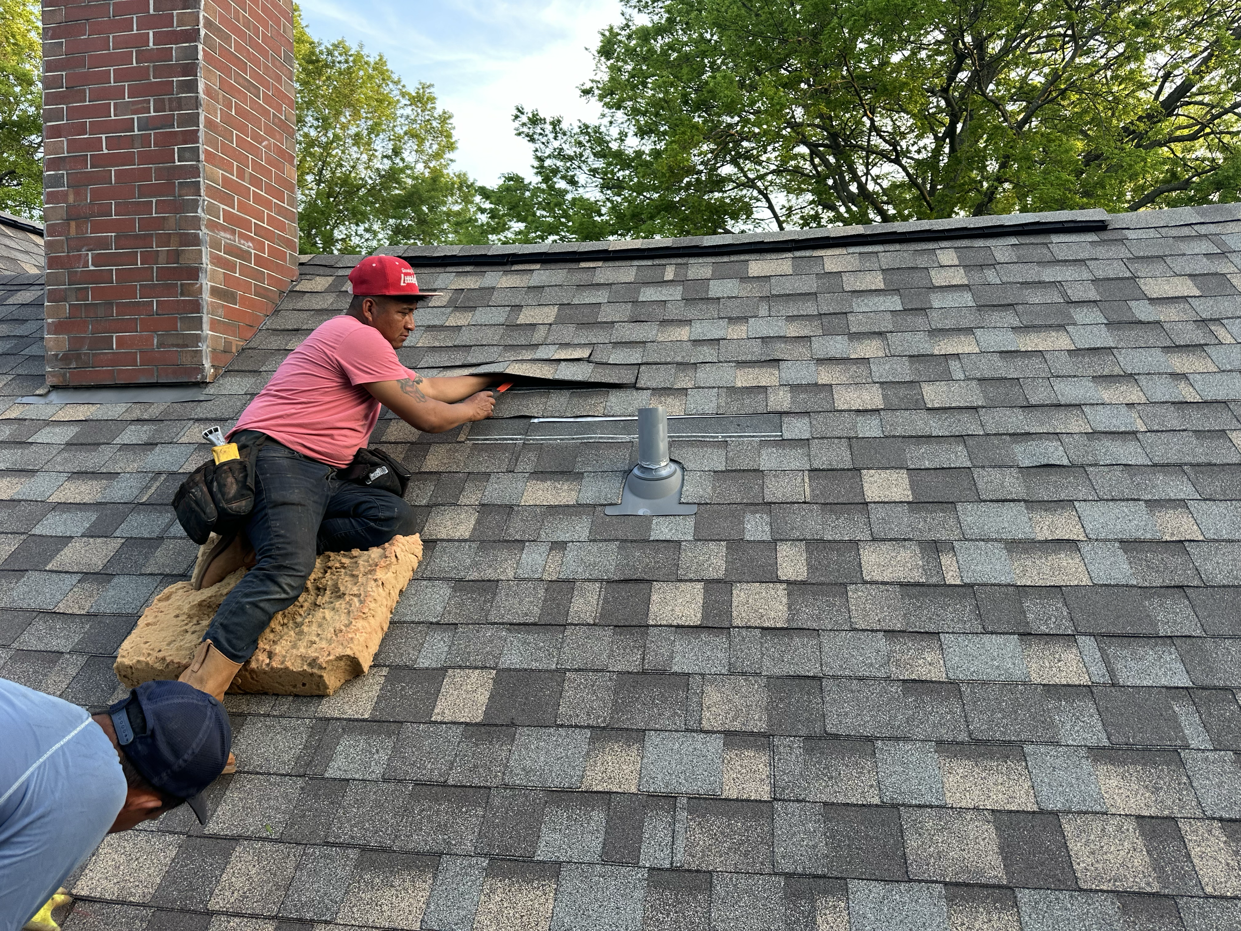 A worker on a roof repairing shingles, with a brick chimney to his side, surrounded by green trees and a clear sky.