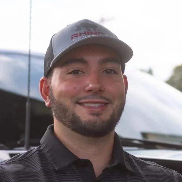 A man outdoors wearing a gray baseball cap with red lettering and a dark polo shirt, smiling with a vehicle and cloudy sky in the background.