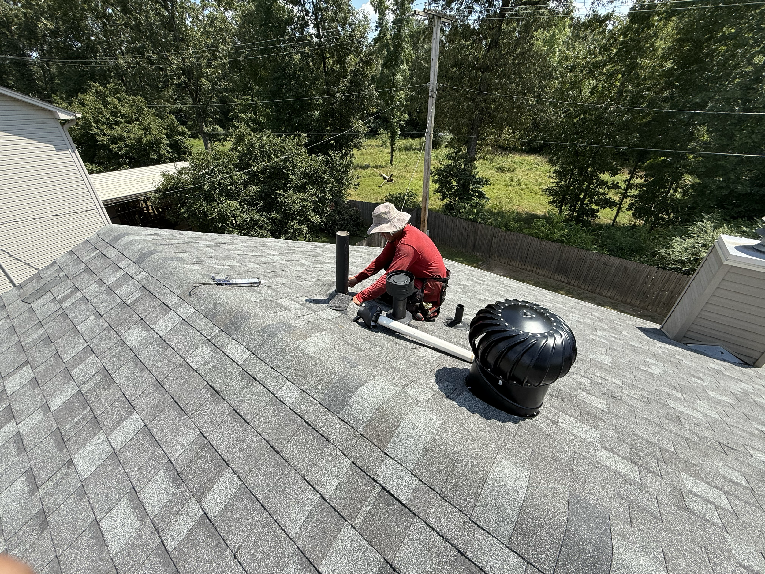A person with a hat working on a roof, inspecting or repairing the vent pipes and other components. The roof is covered with gray shingles, and there's a fenced backyard with trees in the background.