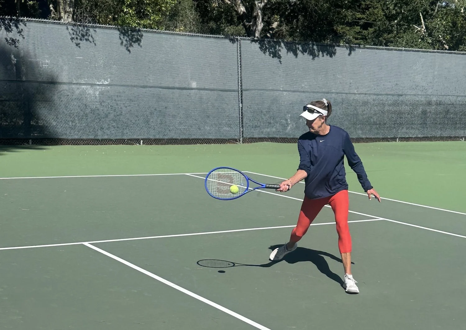 Shelly playing tennis on an outdoor court, wearing red pants, a navy blue long-sleeve shirt, a white visor, and sunglasses, about to hit a tennis ball with a blue tennis racket.