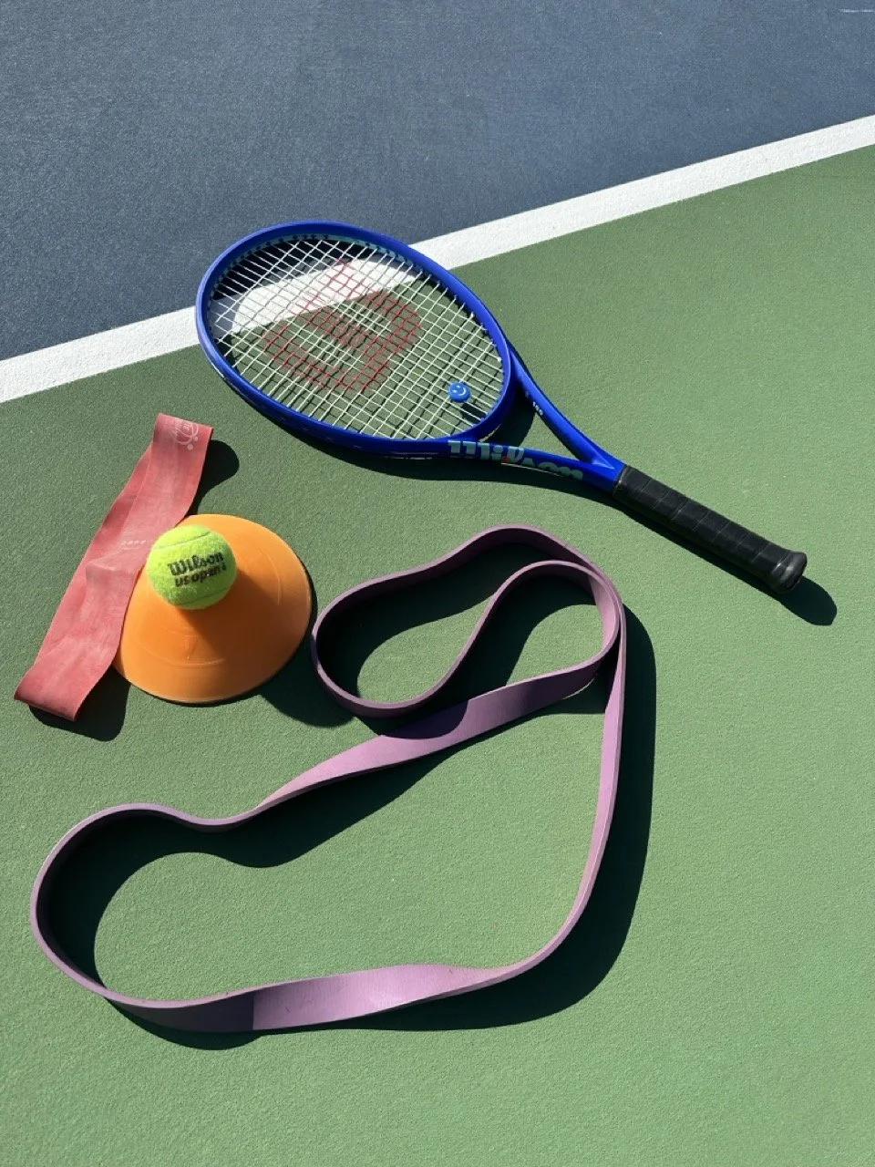 Tennis and sports training equipment including a tennis racket, a tennis ball, an orange cone, and two types of resistance bands on a green and blue tennis court.