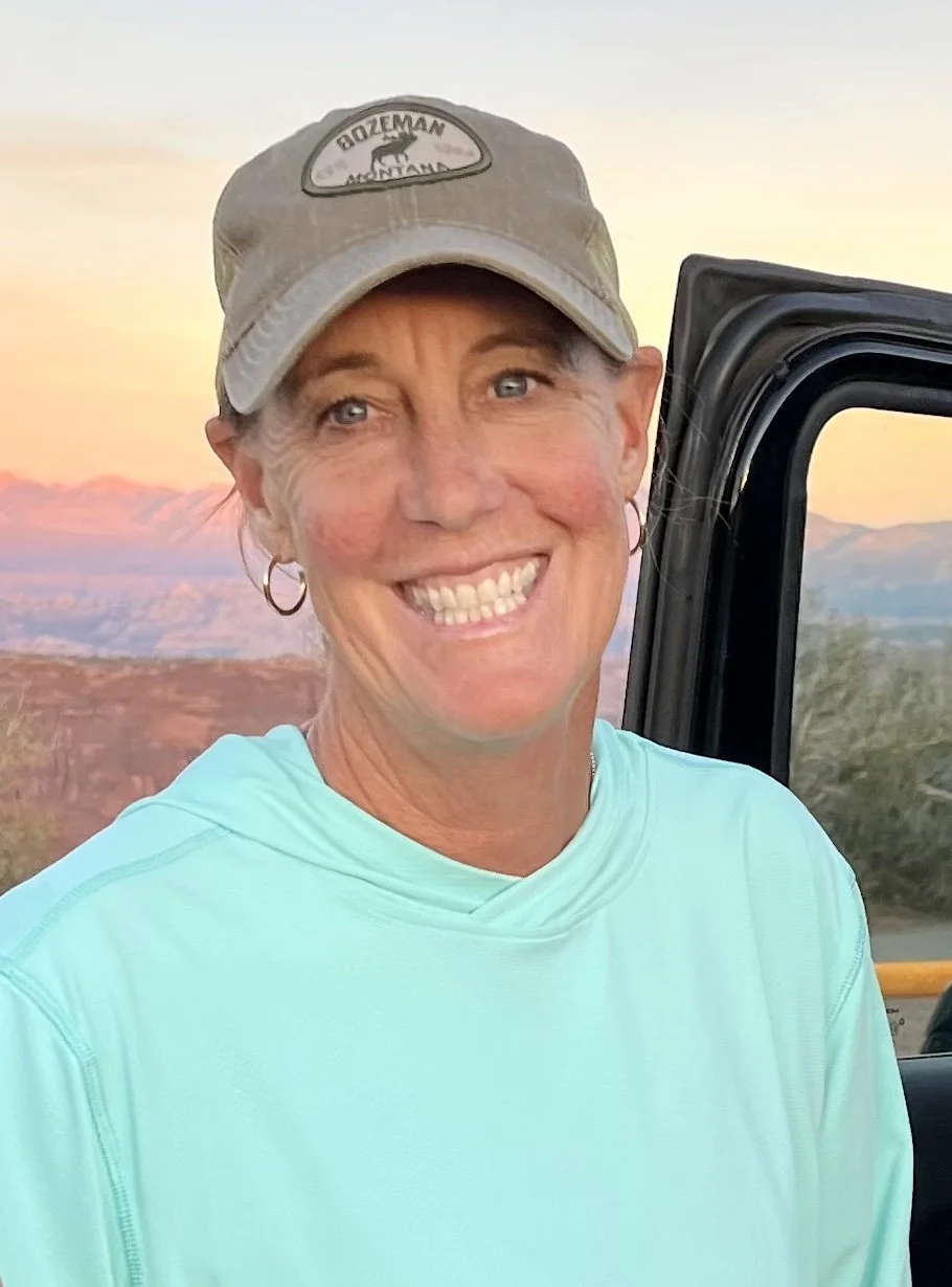 A smiling woman wearing a light blue shirt and a gray cap with a 'Bozeman Montana' patch, standing in front of a landscape at sunset.