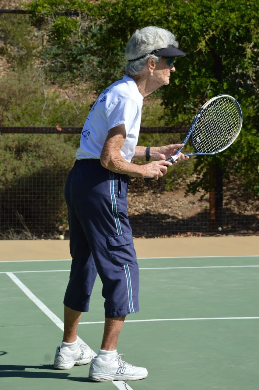An elderly woman with gray hair, wearing sunglasses, a white t-shirt, navy shorts, white athletic shoes, and a black visor, holding a tennis racket on a tennis court outdoors.