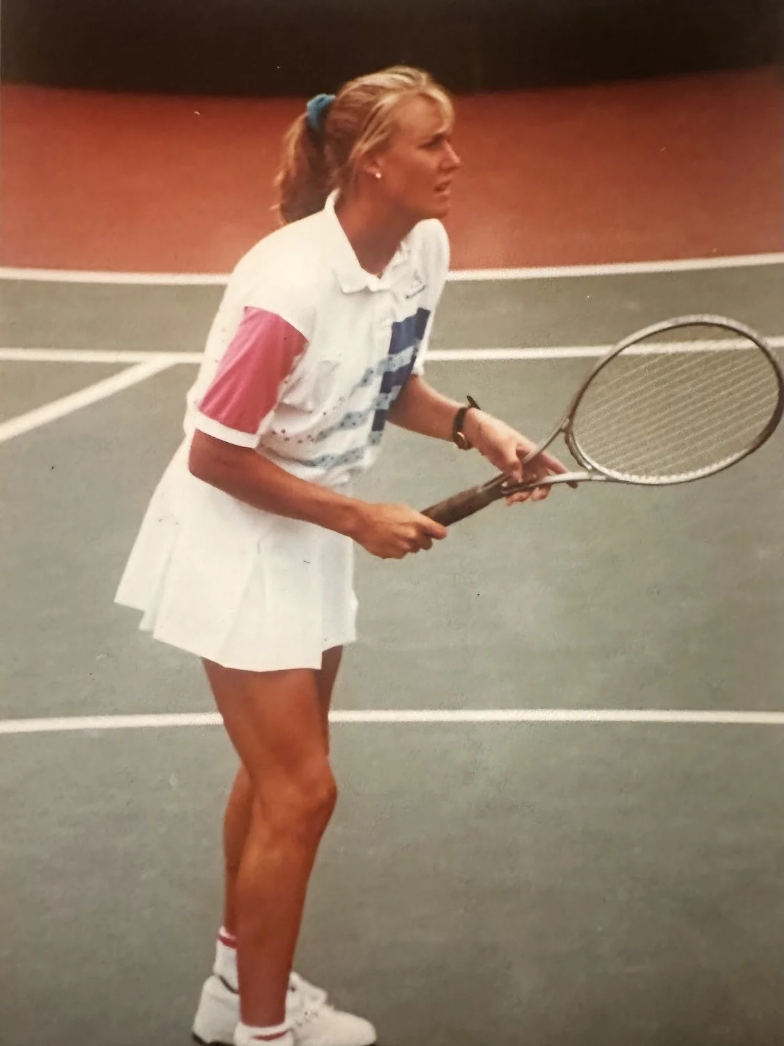 A young Shelly Sweeney on a tennis court holding a tennis racket, dressed in tennis attire with a white skirt and shirt, and a scrunchie in her hair.