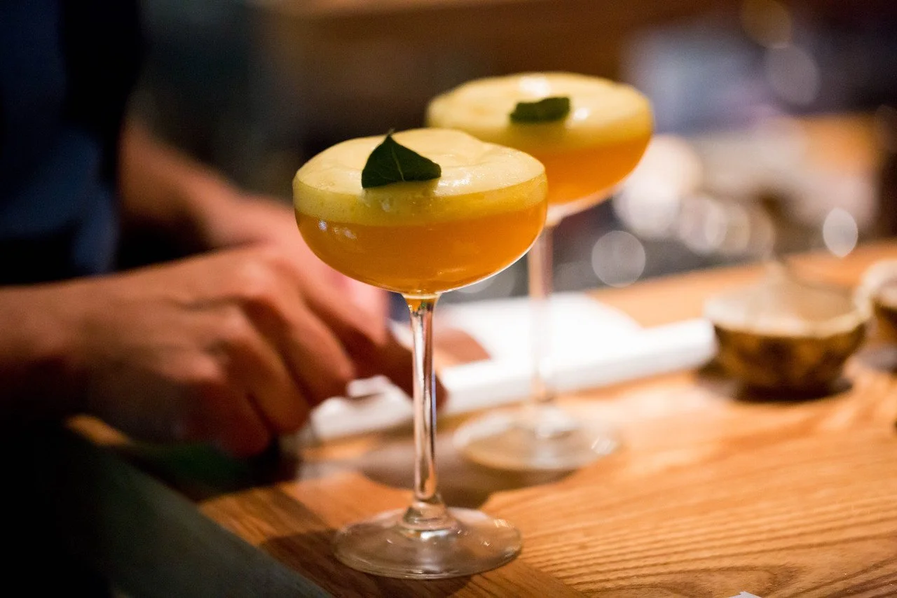 Two cocktail glasses with yellowish drink and green garnish on top, placed on a wooden surface.