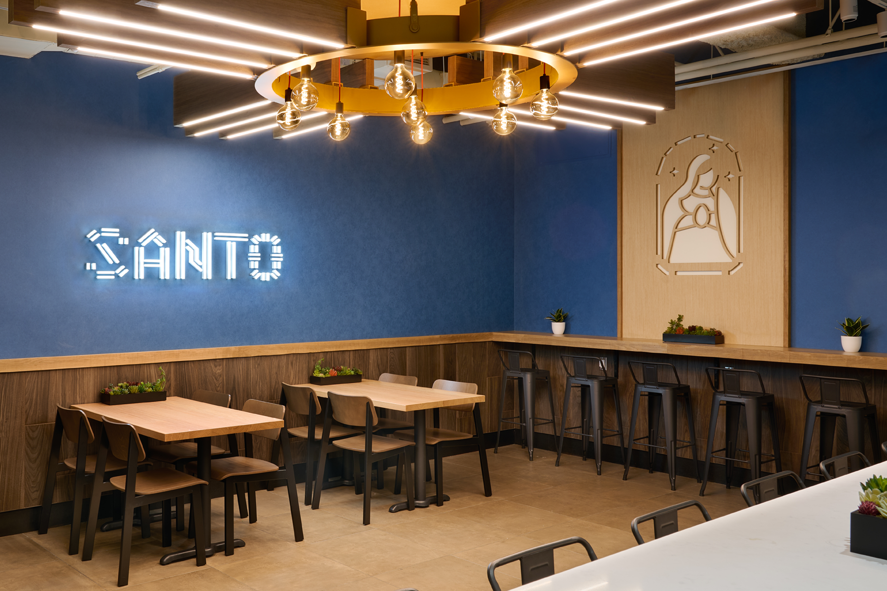 Modern cafe interior with blue walls, wooden furniture, and decorative lighting fixtures. A neon sign reads 'SANTO' on the wall, with a religious-themed artwork of a woman in a frame. Small potted plants adorn the tables and counter.