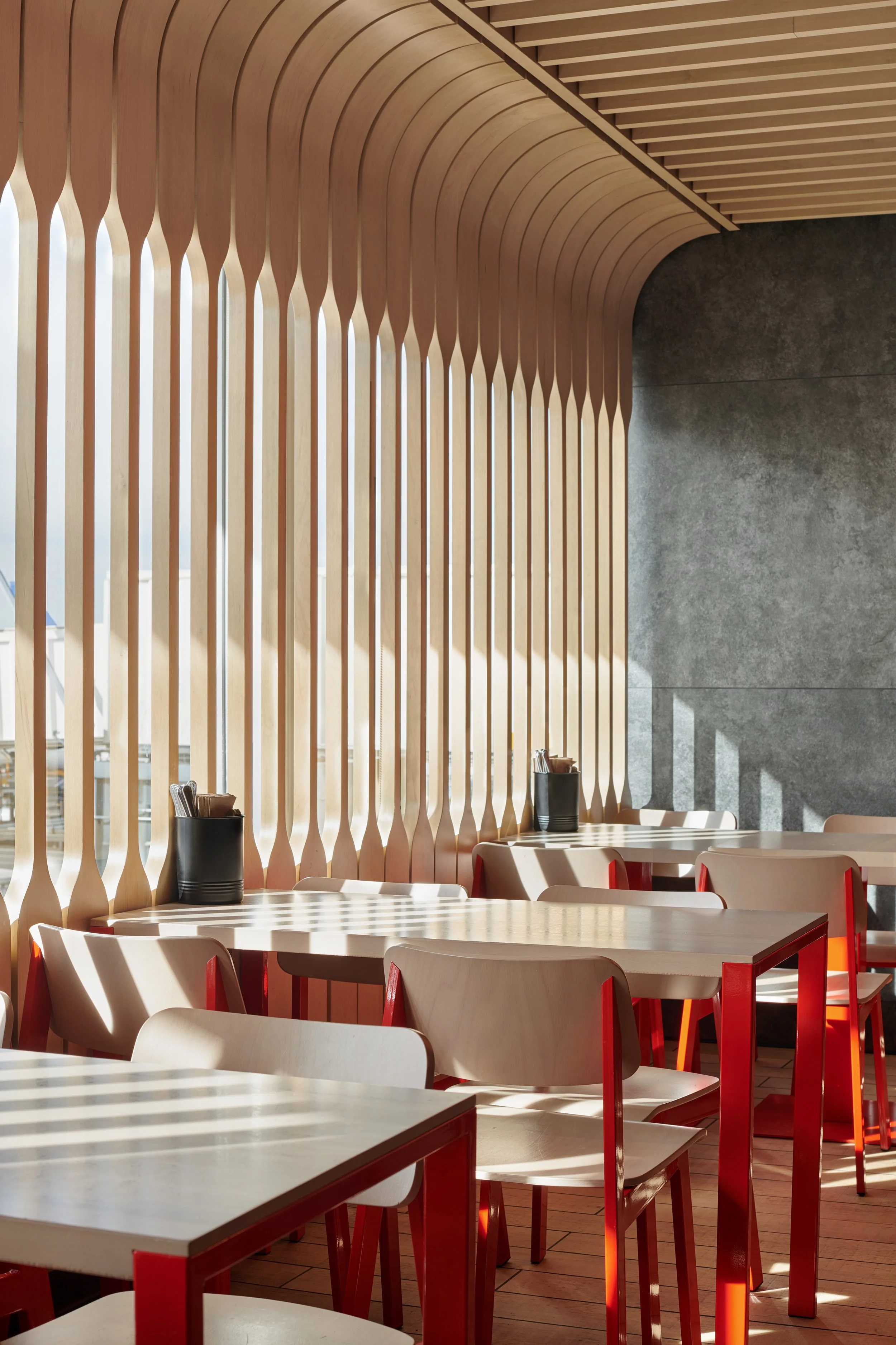 Interior of a modern restaurant with wooden partitions and white tables with red chair frames, illuminated by sunlight.