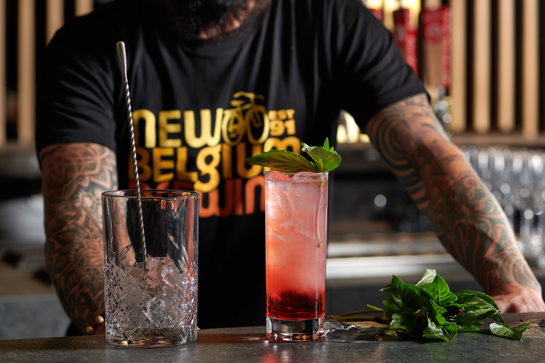 A bartender creates a pink cocktail garnished with green leaves, sitting on a bar counter with an empty glass filled with ice and a black and silver drinking straw beside it. The bartender's forearms are tattooed, and they are wearing a black T-shirt with yellow and orange text that reads ‘New Belgium’ and other text. A bunch of green leaves is on the bar in front of the cocktail.
