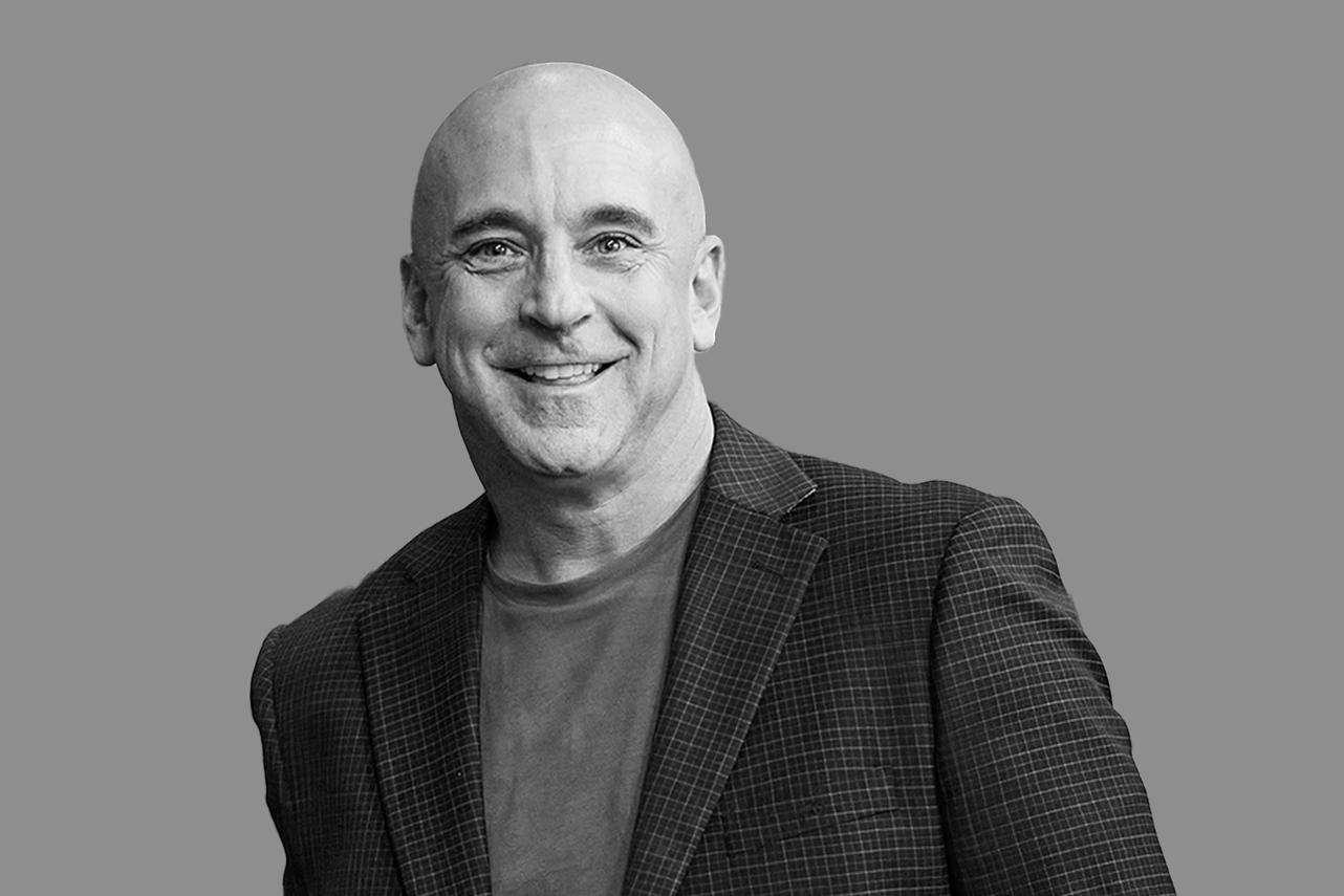 Black and white portrait of a smiling bald man wearing a checkered blazer and T-shirt, against a plain gray background.