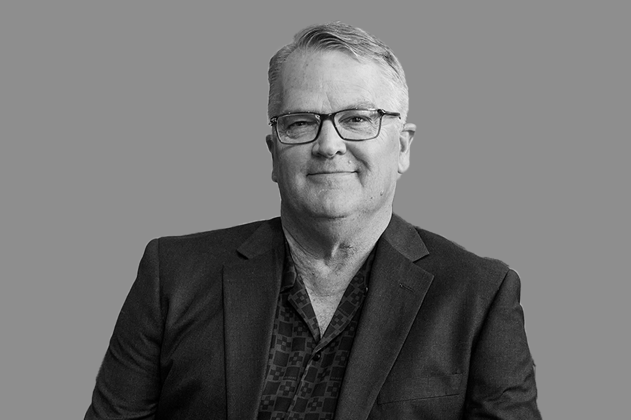Black and white portrait of a middle-aged man with glasses, dressed in a dark blazer and patterned shirt, smiling slightly against a plain background.