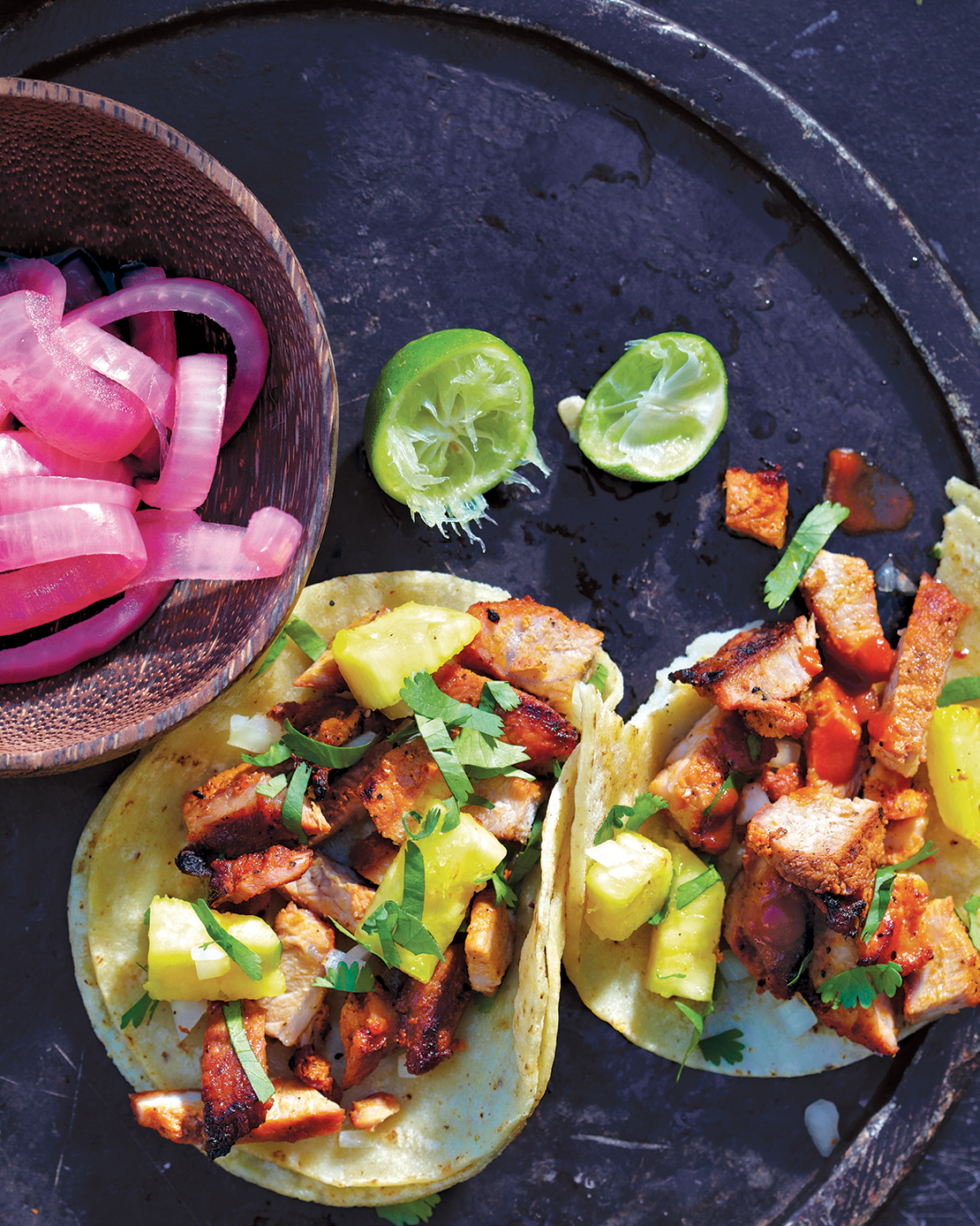 Two grilled tacos filled with chopped grilled chicken, pineapple, and cilantro, accompanied by chopped onions, sliced jalapeños, and lime wedges on a black platter.