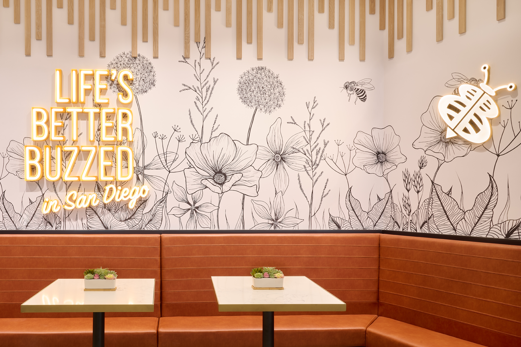 Decorative wall with black floral line art, illuminated neon sign reading "Life's Better Buzzed in San Diego," and a stylized neon bee on the wall. There are two square tables with small flower arrangements and a brown leather bench seat along the wall.