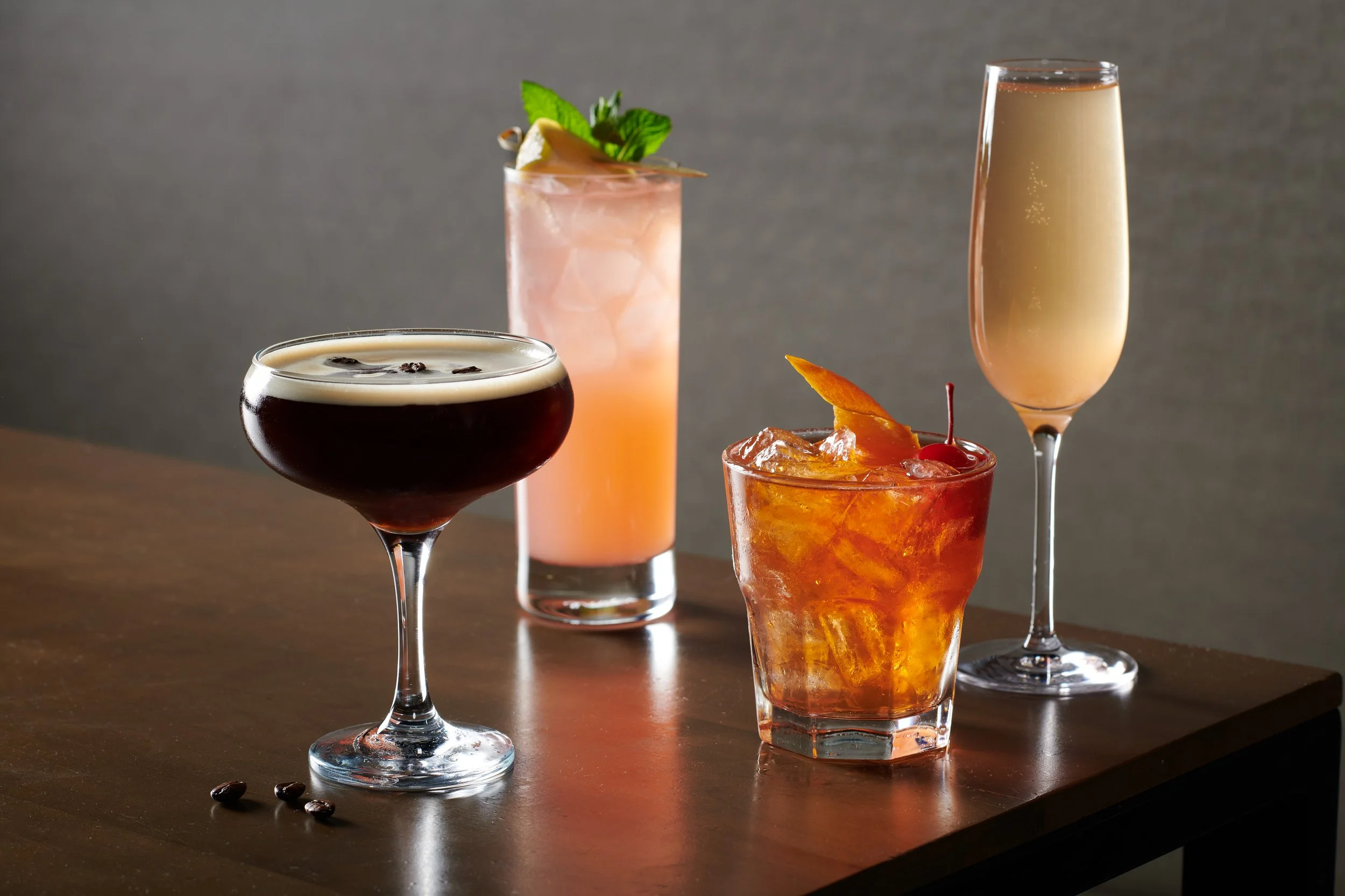 Four different cocktails on a wooden surface, with a gray background, including a dark cocktail with a foamy top, a pink drink with mint garnish, an amber-colored drink with a cherry and orange peel, and a pale yellow champagne in a flute glass.