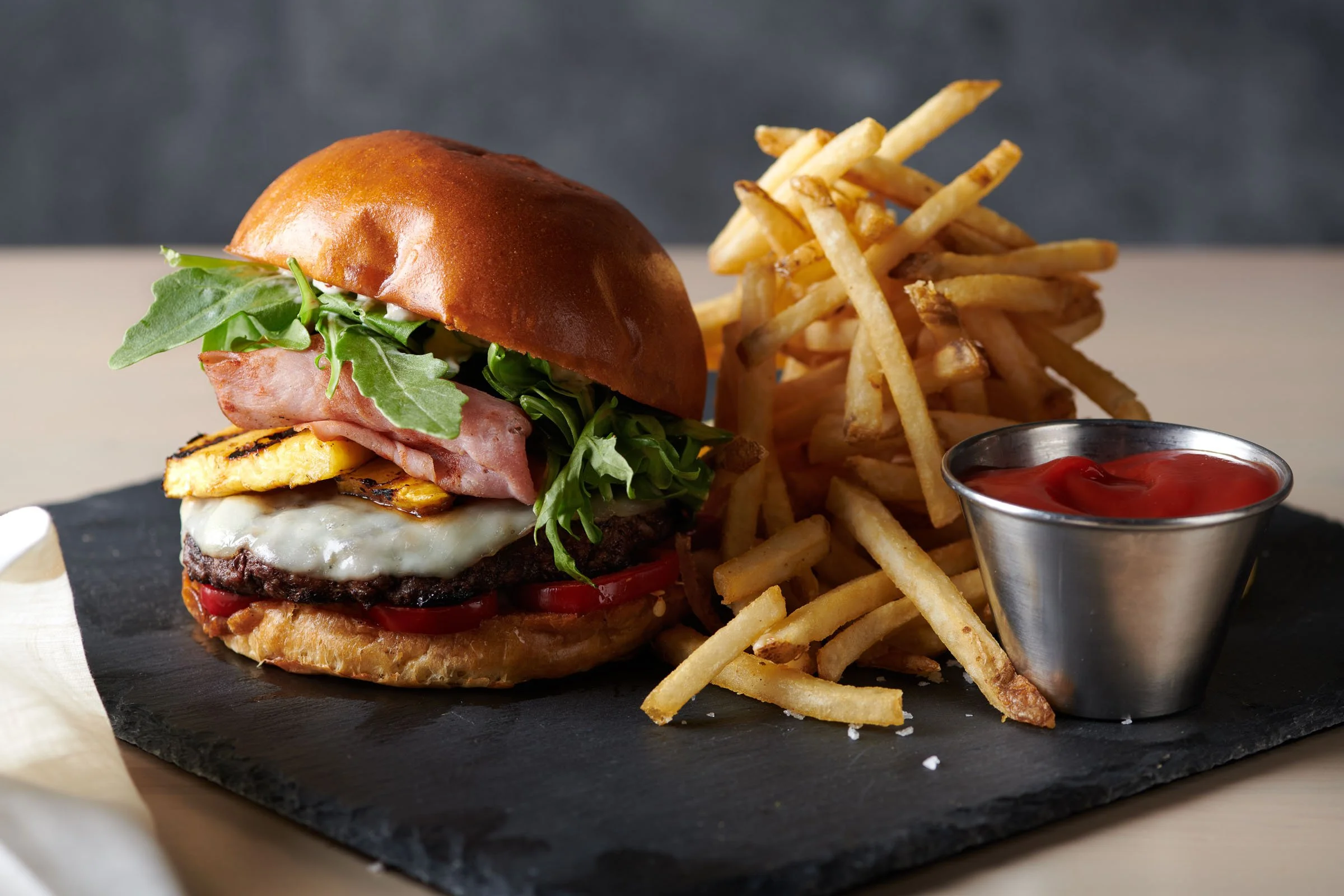 A gourmet hamburger with a brioche bun, lettuce, tomato, a fried egg, cheese, ham, and a beef patty, served with French fries and a small cup of ketchup on a black slate serving board.