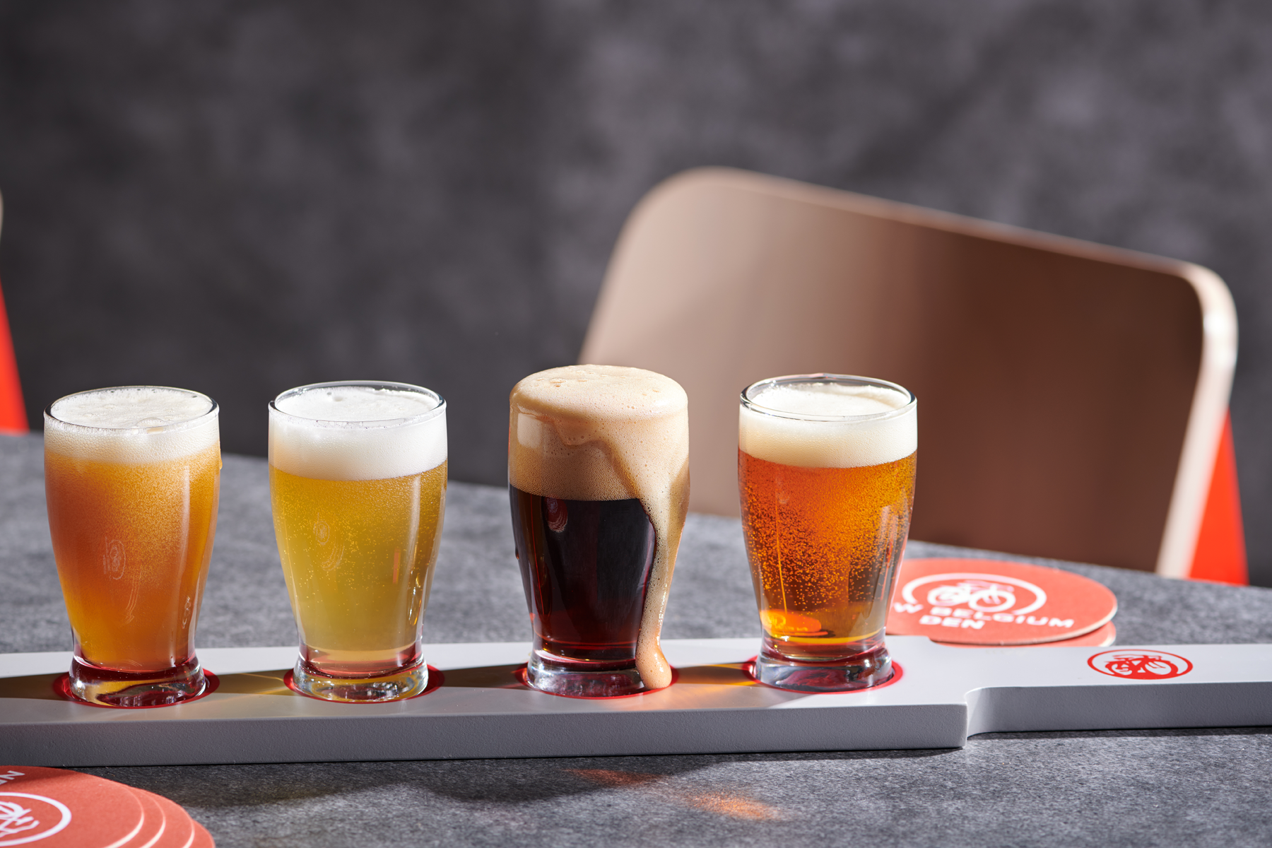A beer flight with four different beers on a gray table, with a background of a gray wall and beige chair.