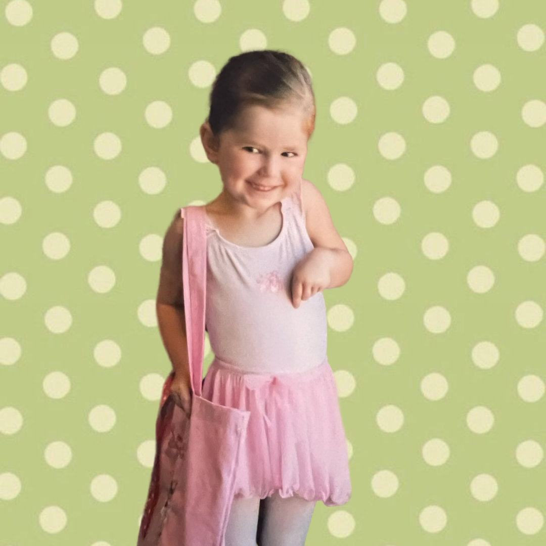 A young girl with dark hair in a bun, wearing a pink tutu dress, gray tights, and carrying a pink bag, standing against a light green background with white polka dots, smiling and pointing at herself.