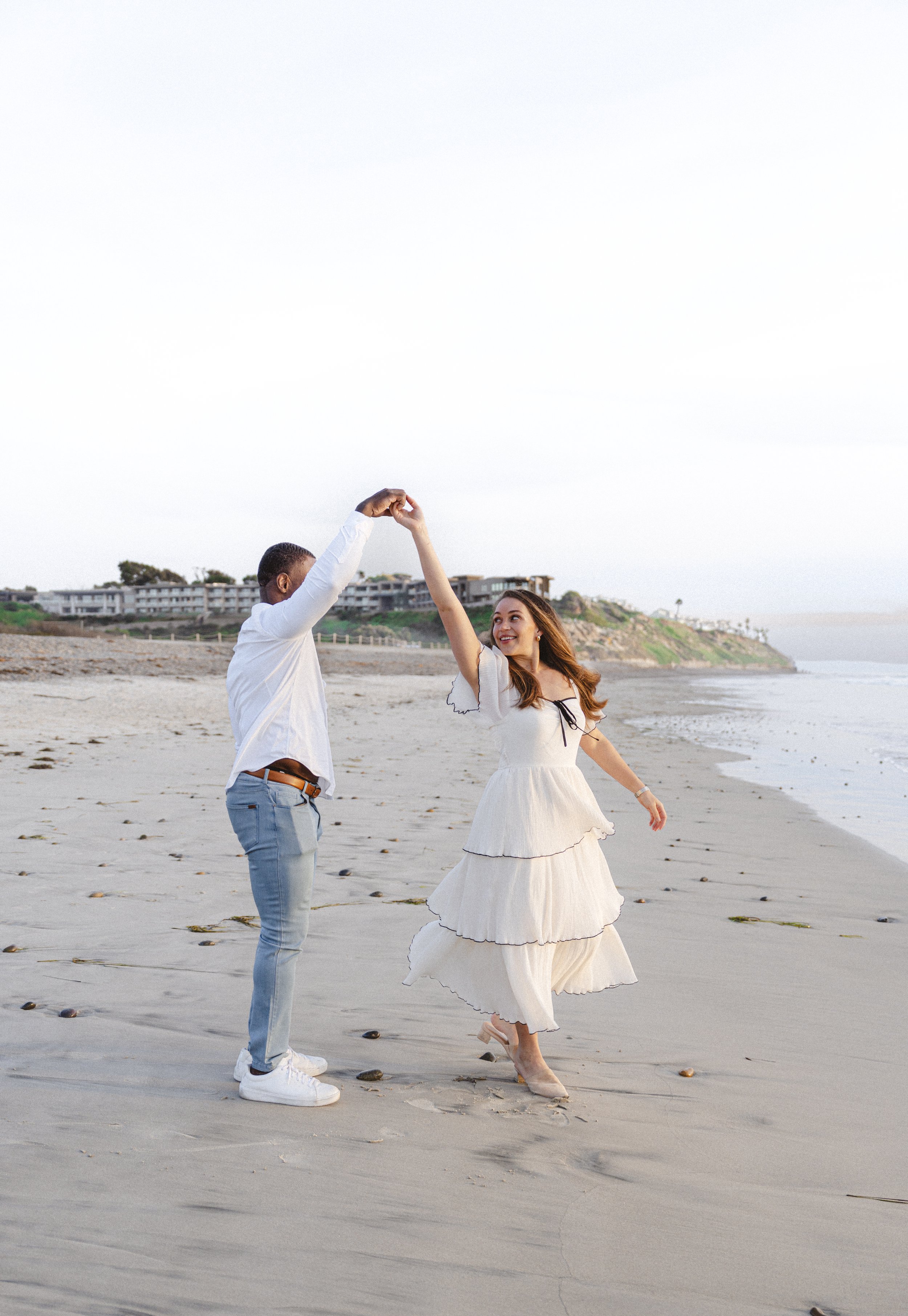Couple twirling and smiling on the sand during a light-filled couples photoshoot at Ponto Beach in Carlsbad, California.